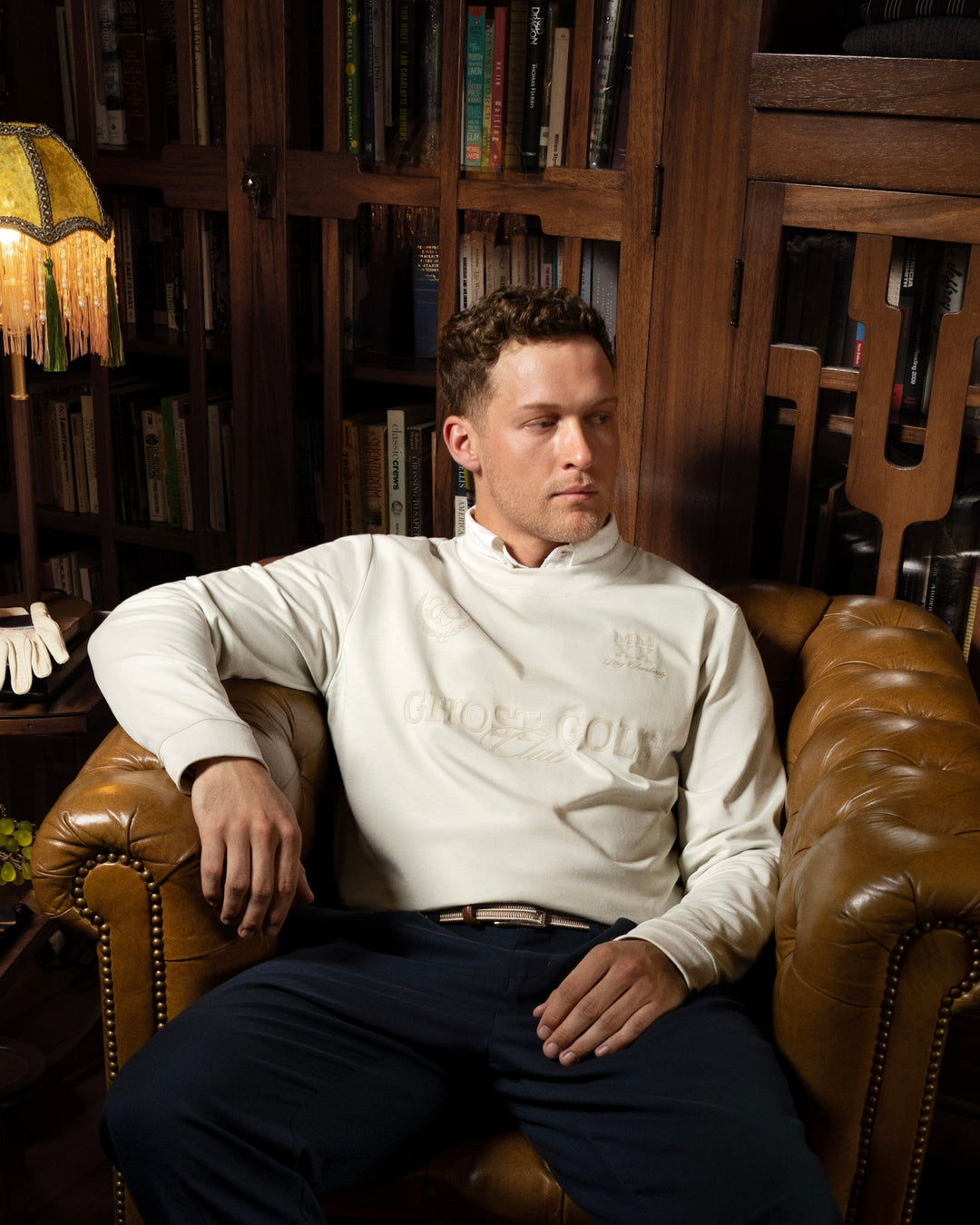 Lifestyle image of a male model sitting in a vintage-style chair inside a vintage room, with one arm resting on the armchair and a relaxed, confident expression.