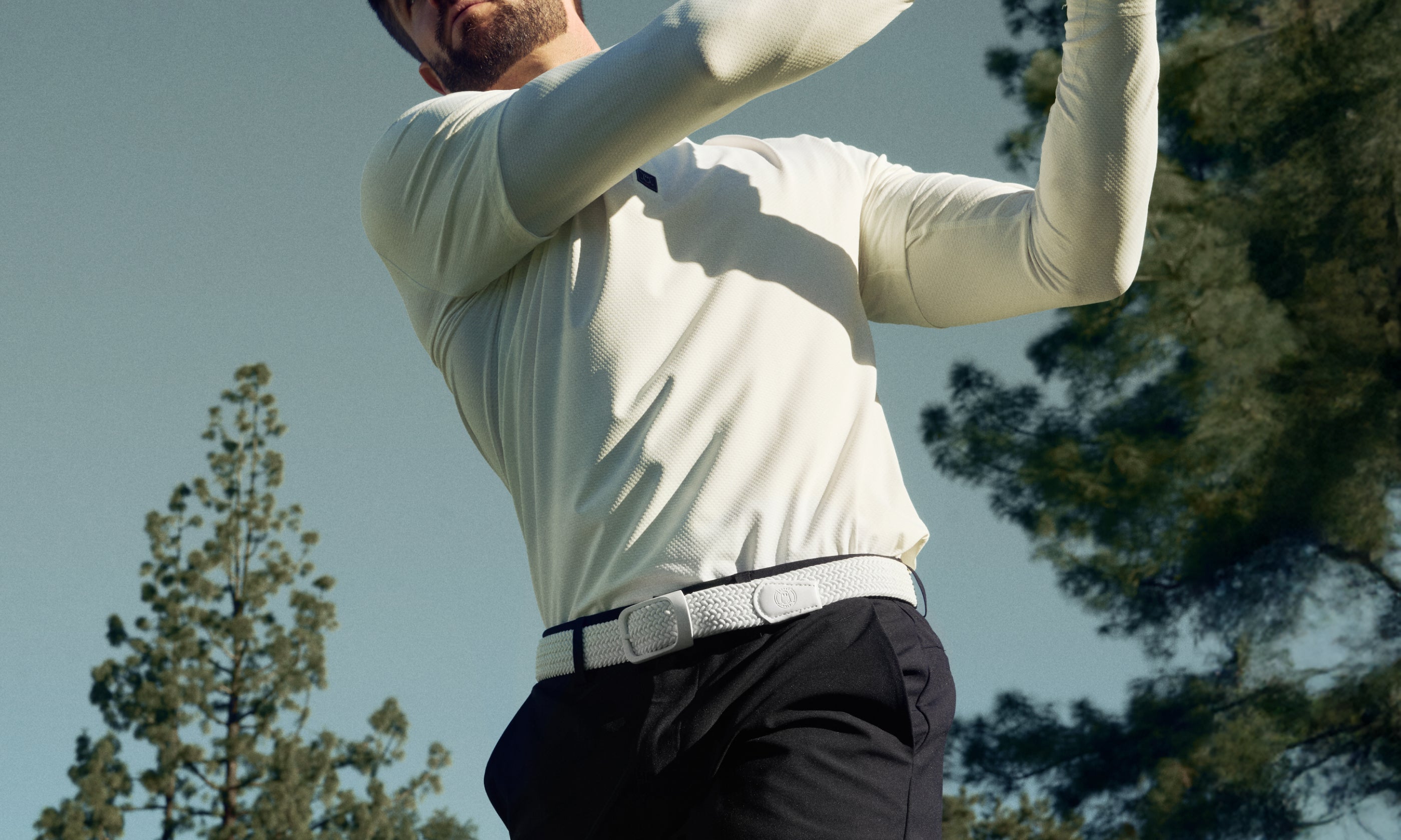Man swinging a Golf Club wearing a White Polo and Black Shorts with White Belt