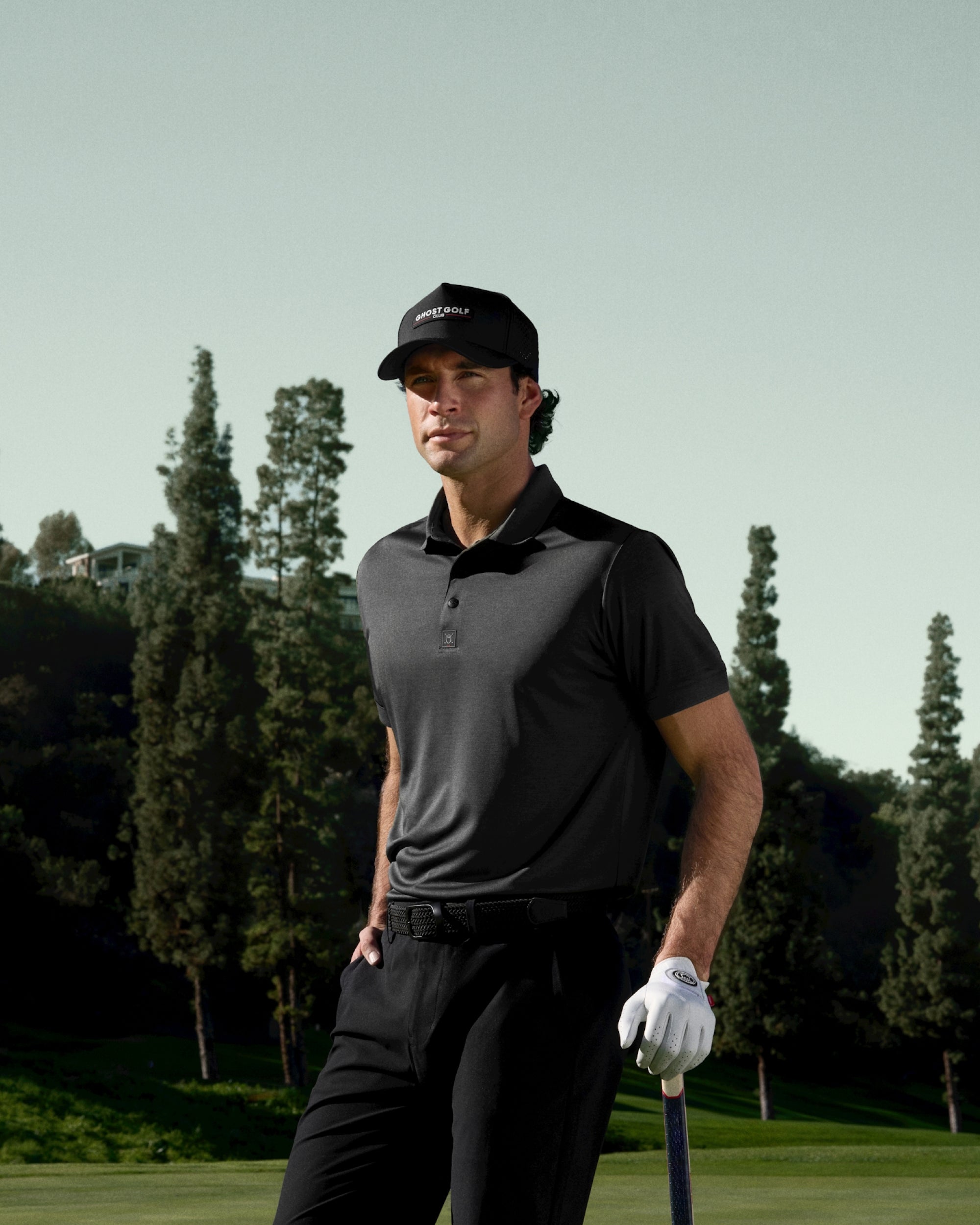 White male golfer on a golf course wearing a grey polo tucked into black pants with a black belt.
