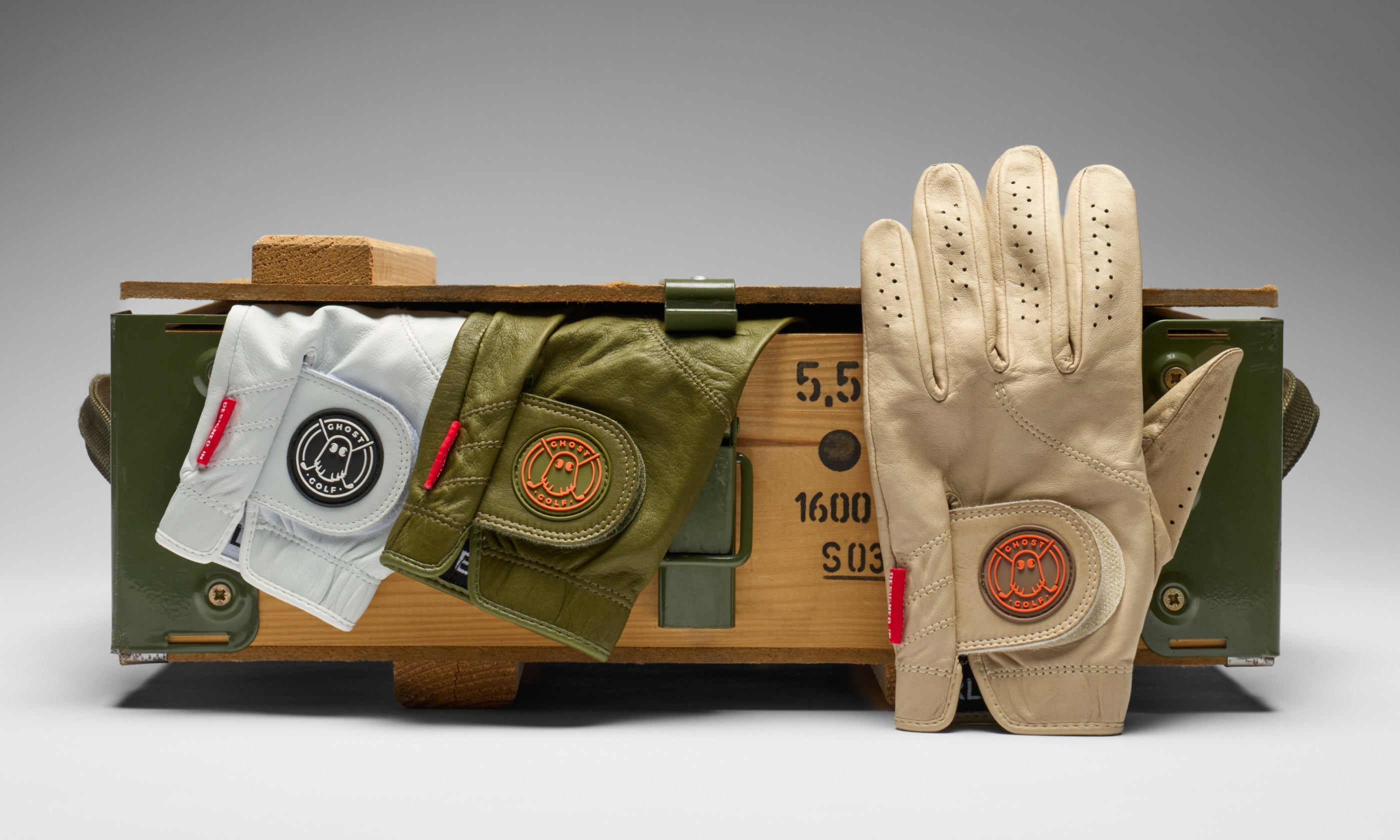 Studio product photo of Ghost Golf gloves, with a pair partially visible inside a small wooden crate and one glove displayed upright. The Ghost Golf logo is prominently featured on the glove.