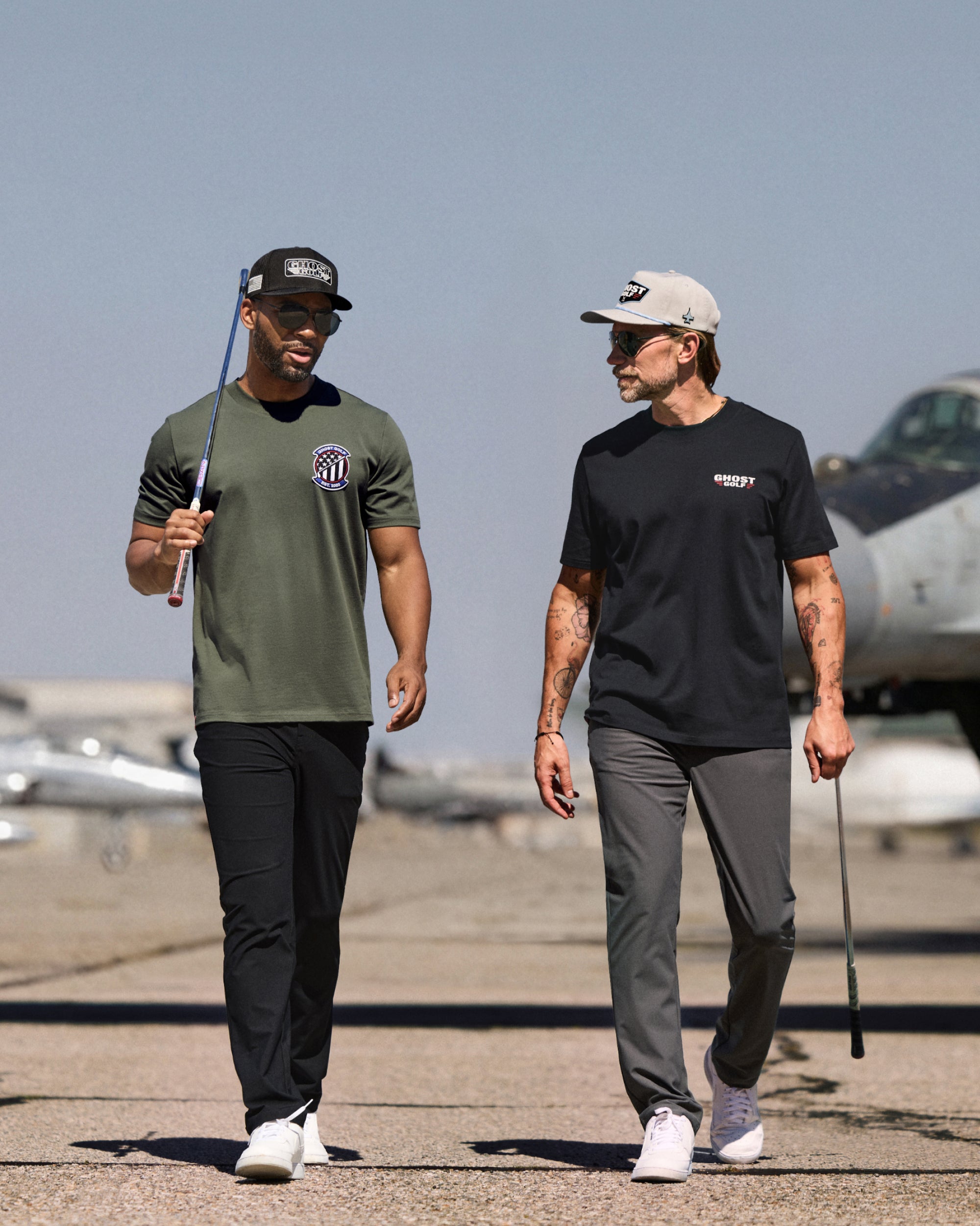 Lifestyle photo of two male models walking toward the camera while holding golf clubs. Both are dressed in golf apparel, including T-shirts, hats, and pants. Out-of-focus fighter jets in the background create an aviation-inspired setting, while the models remain the focal point of the image.