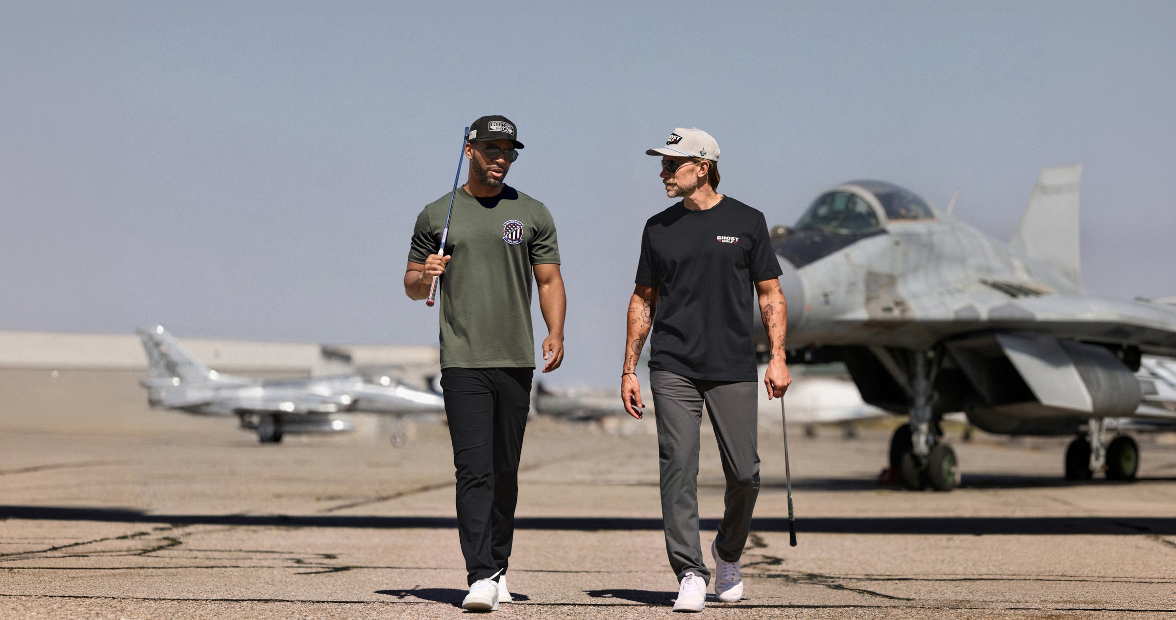 Lifestyle photo of two male models walking toward the camera while holding golf clubs. Both are dressed in golf apparel, including T-shirts, hats, and pants. Out-of-focus fighter jets in the background create an aviation-inspired setting, while the models remain the focal point of the image.