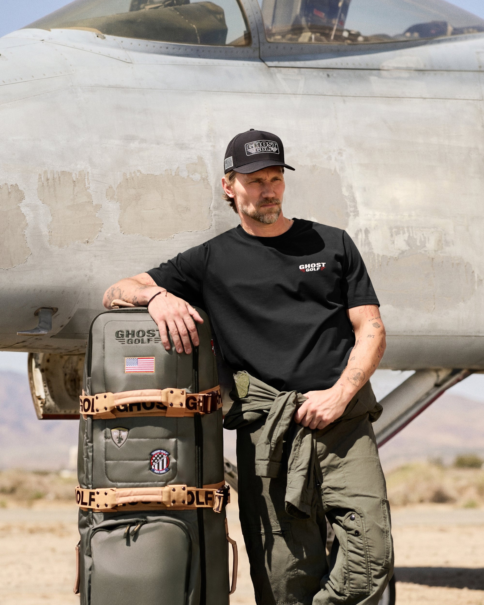 Lifestyle image of a white male model standing in front of a large military-style aircraft, looking to his left. He is leaning his right arm on a travel golf bag.