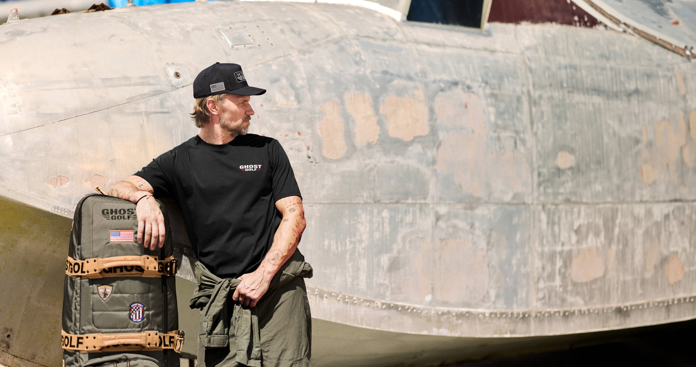 Lifestyle image of a white male model standing in front of a large military-style aircraft, looking to his left. He is leaning his right arm on a travel golf bag.