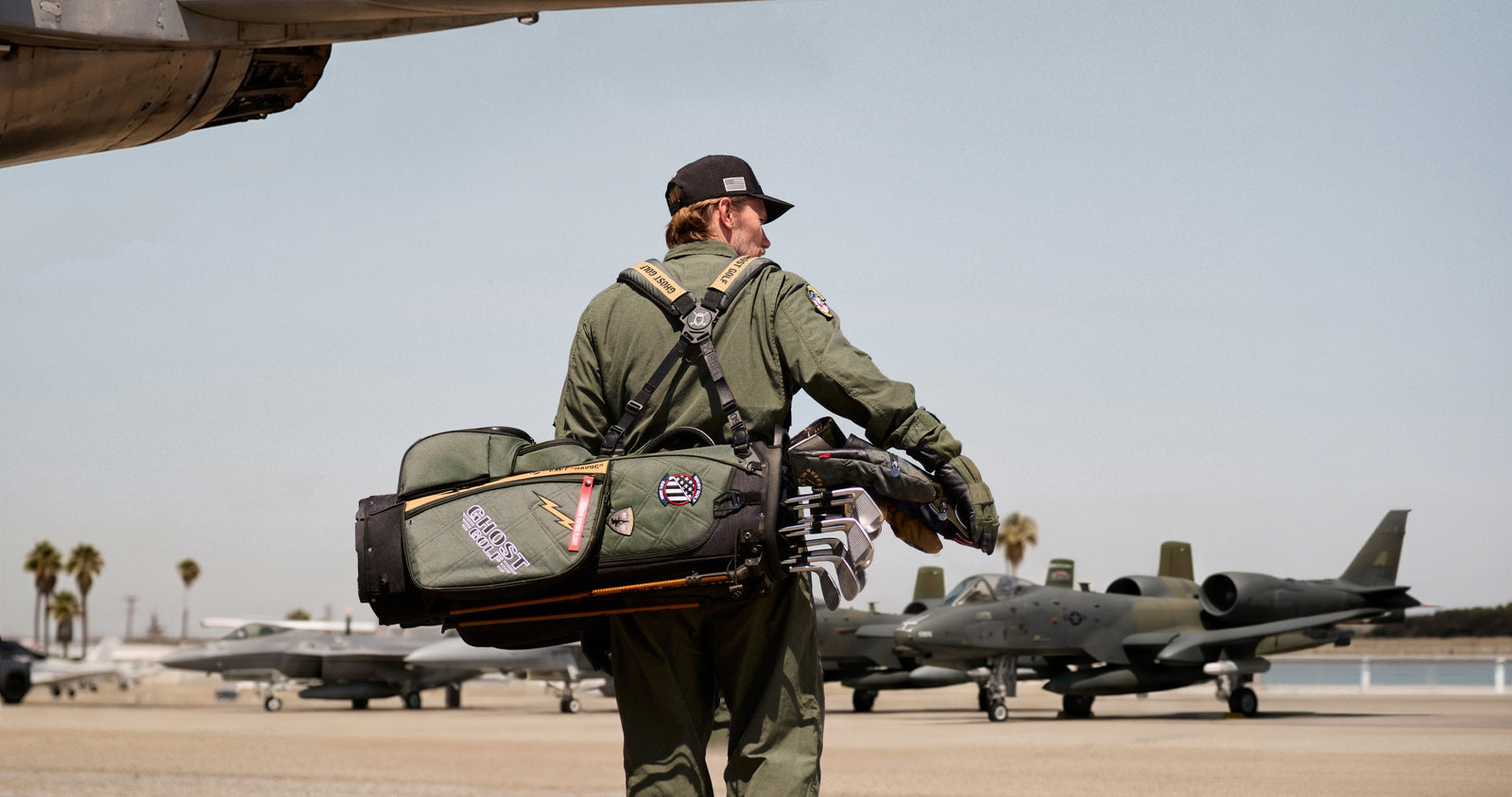 Lifestyle photo of a male model carrying a G-Suit Anyday Golf Bag in an aviation-inspired setting. Fighter jets are visible in the background, creating the impression of a military airfield while keeping the focus on the model and travel-ready golf bag.