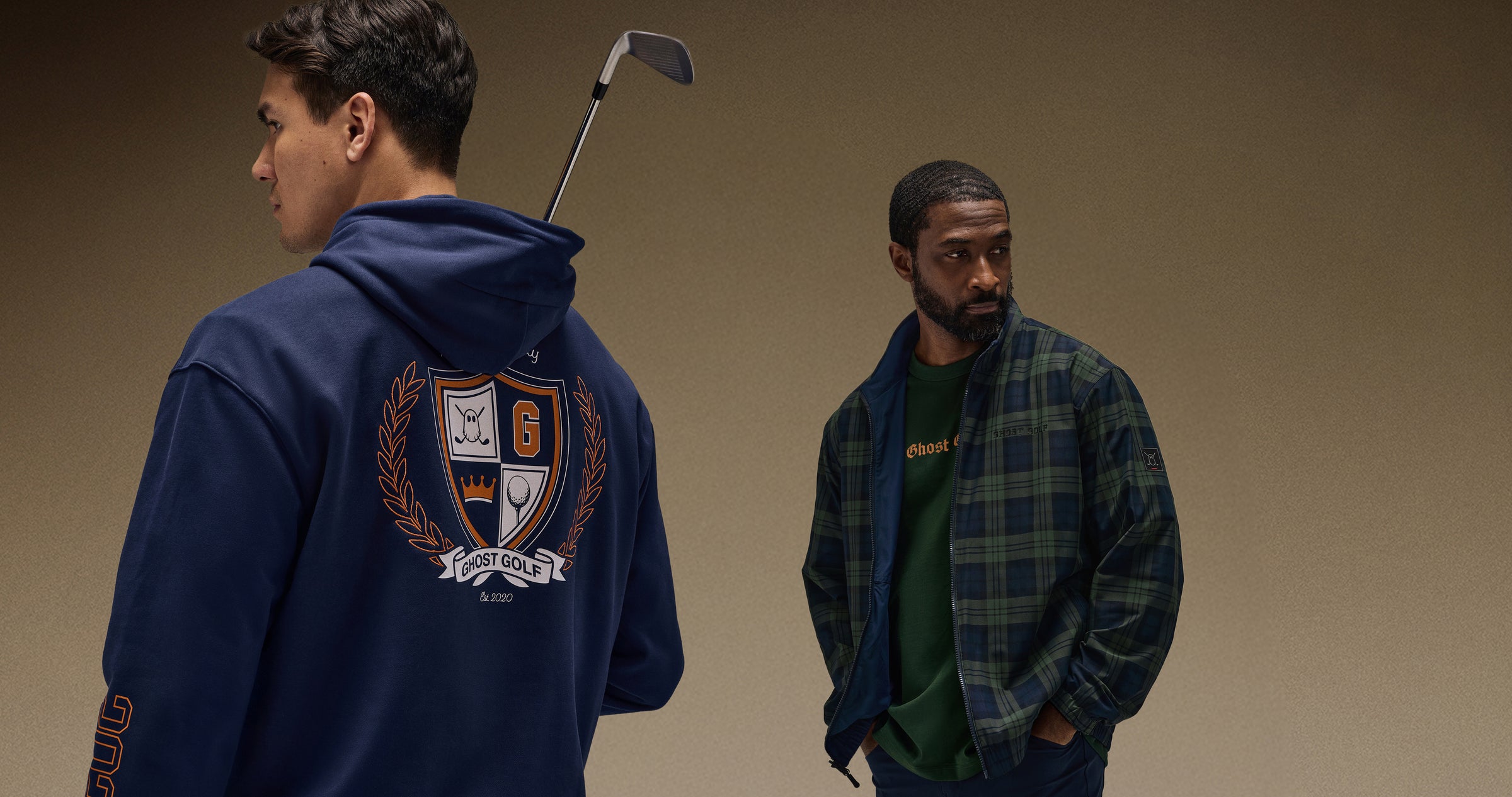Asian golfer in brown studio, back facing, in a navy hoodie with back graphic, holding a golf club. Black golfer wears reversible plaid navy jacket and green tee, hands in pants.