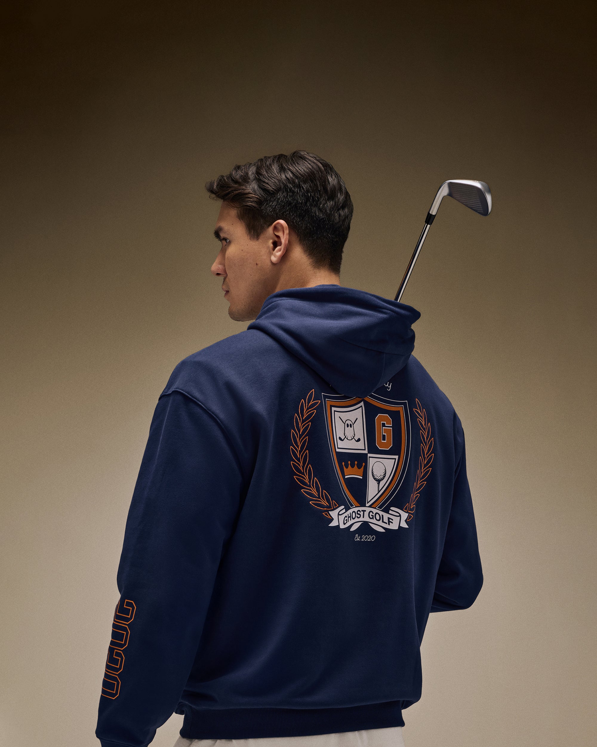 Asian golfer in brown studio, back facing, wearing a navy hoodie with back graphic, holding a golf club.