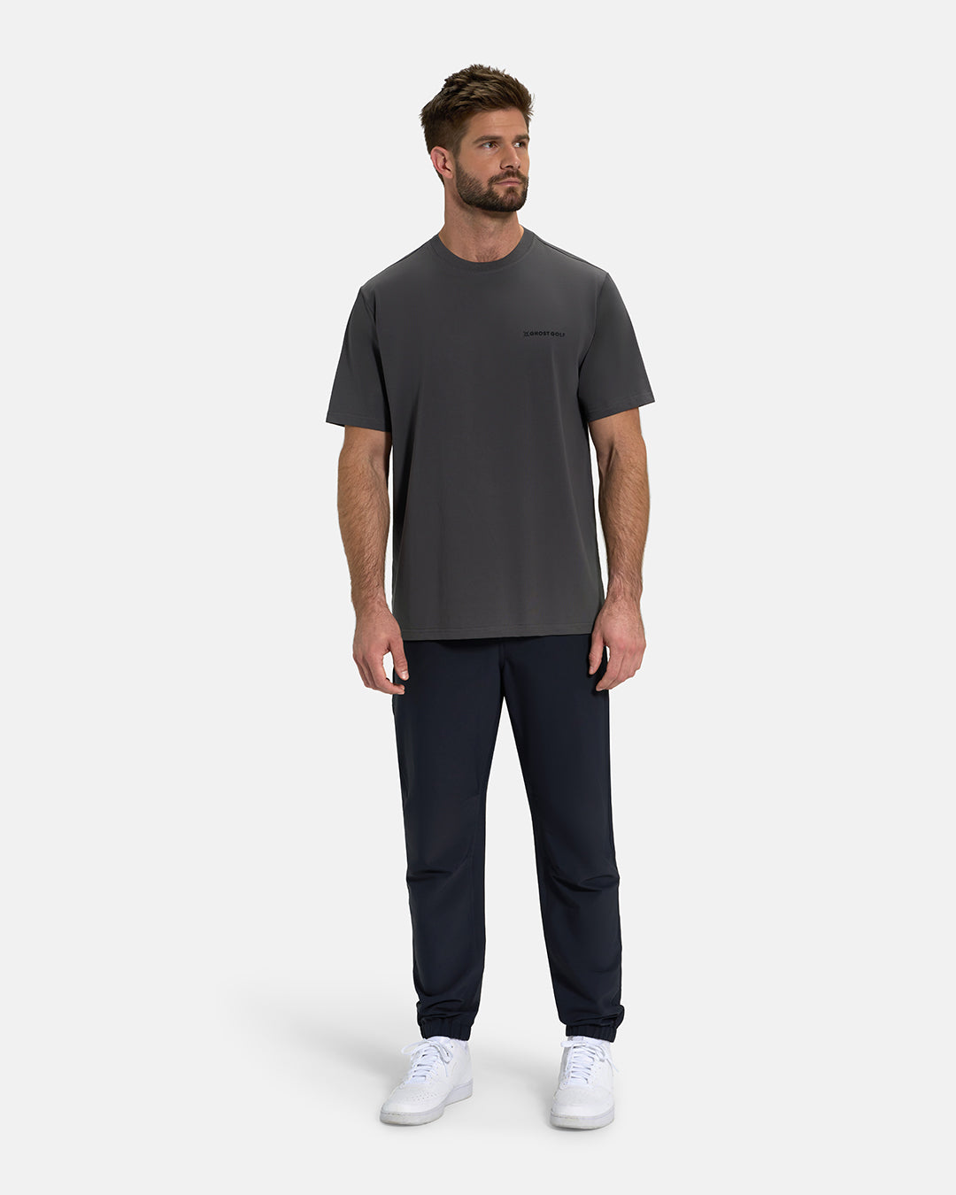 Full Body View of Iron Grey Ghost Golf Core Logo Tee on White Male Model wearing navy pants and white shoes #color_iron-grey