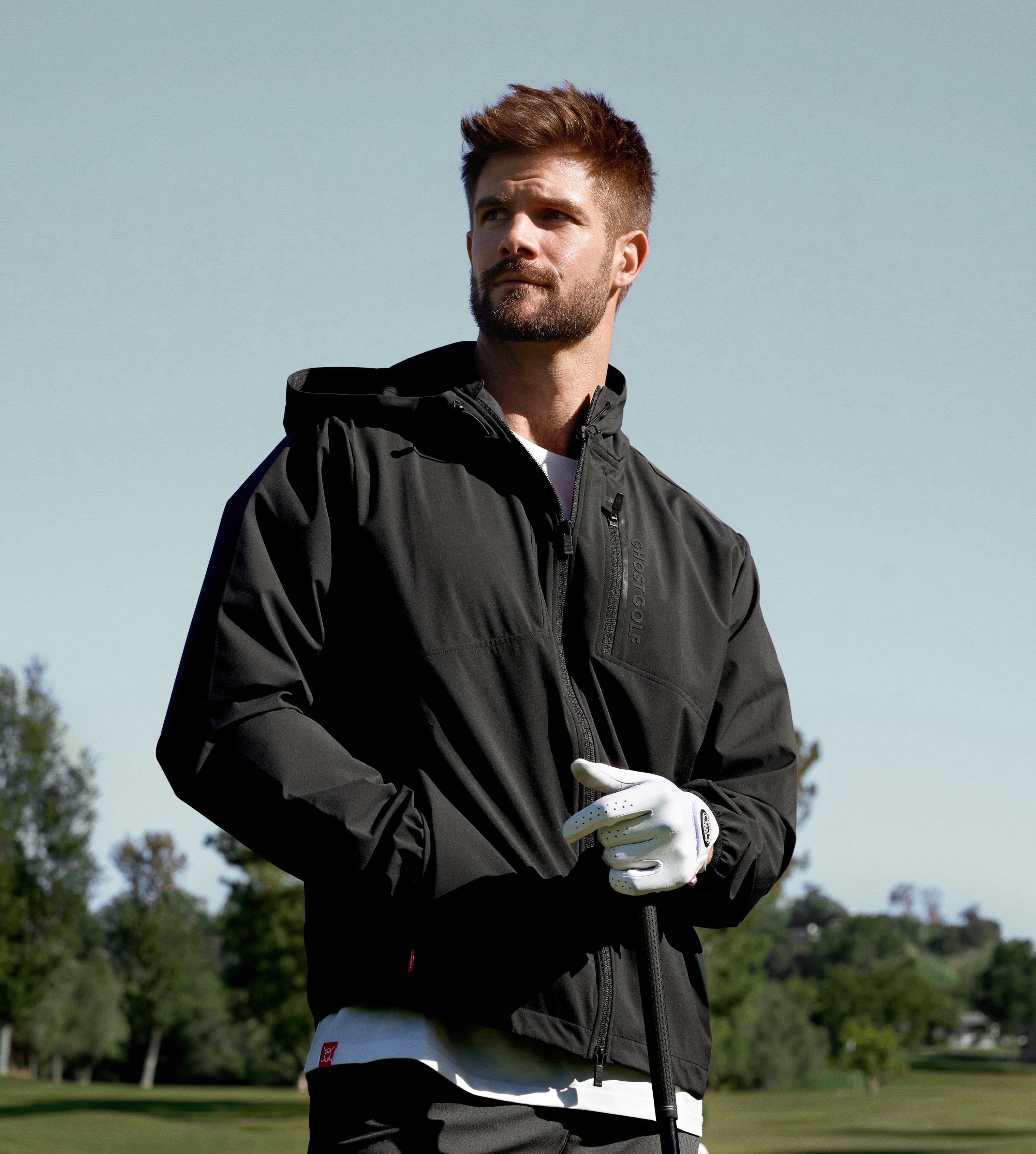 White male golfer on a golf course wearing a black jacket over a white tee, holding a golf club with a white golf glove.