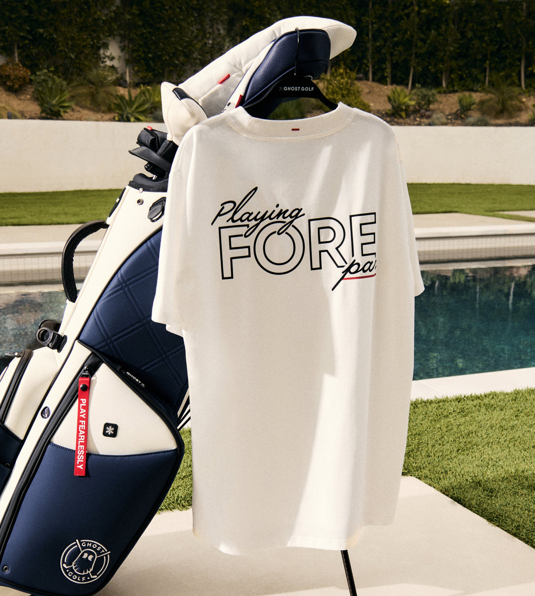 A T-shirt is draped over a golf club headcover on a golf bag near the pool, creating a stylish lifestyle display. The back of the shirt features a graphic with the phrase “Playing Fore Par,” adding a golf-inspired touch to the poolside setting.