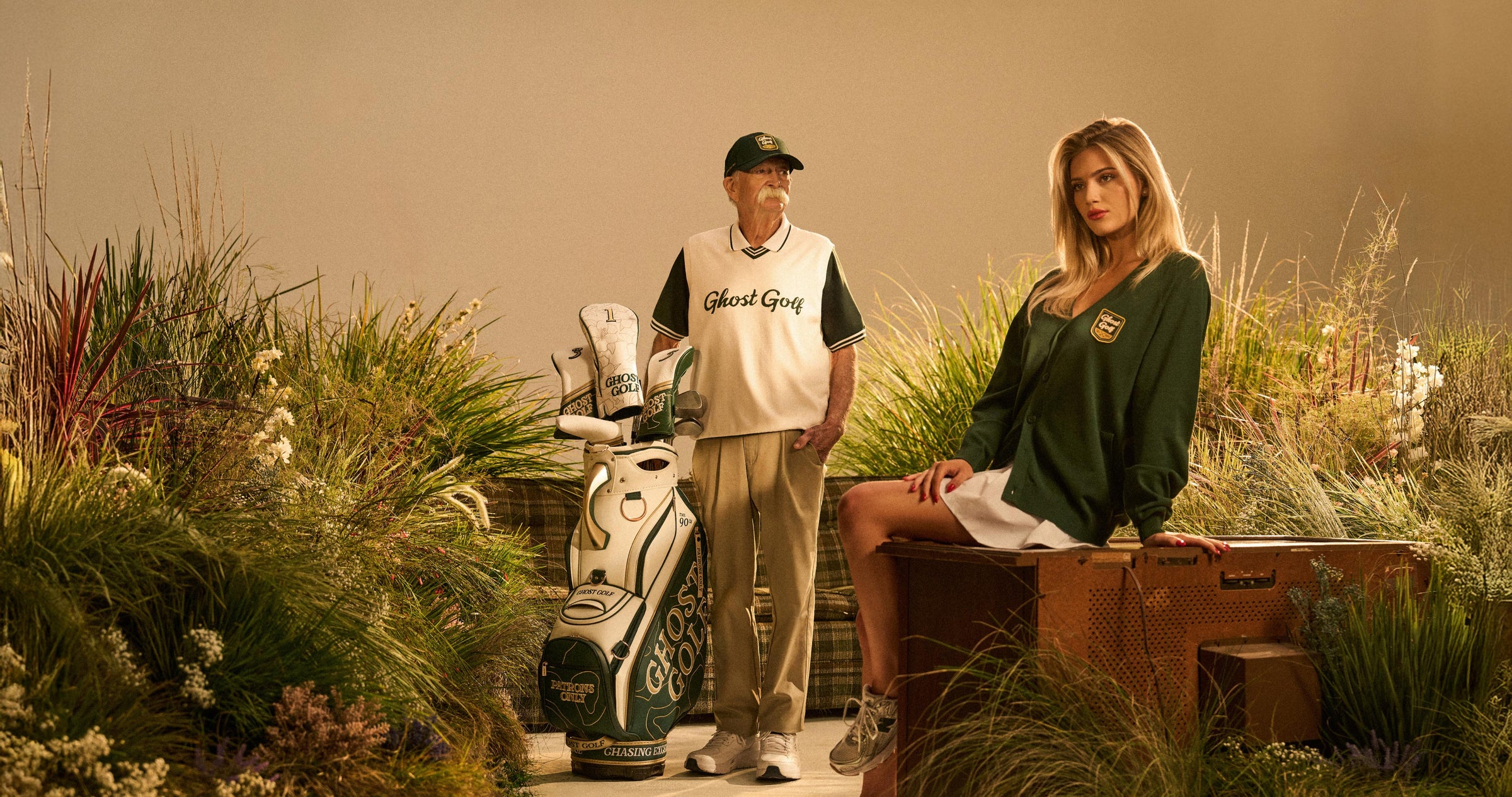 Senior male model holding a golf staff bag in a studio with grass scenery, wearing a green sweater vest, green hat, and khaki pants. A female model sits on a vintage television nearby, wearing a green cardigan and white skirt.
