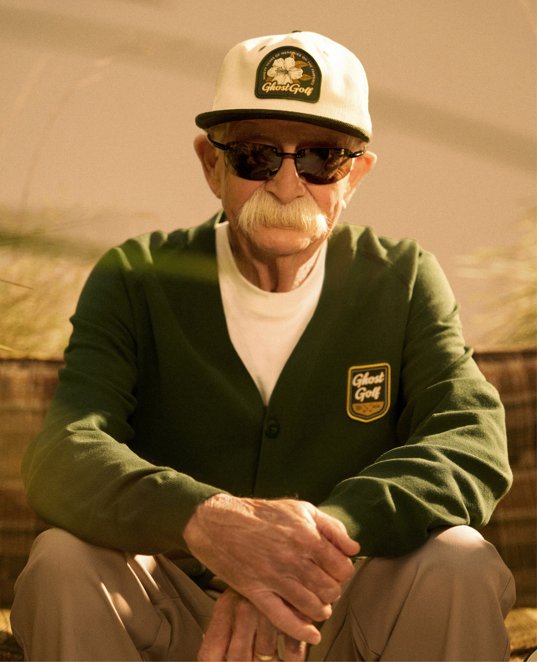 Senior Male Model sitting on a couch wearing green cardigan white hat khaki pants and glasses