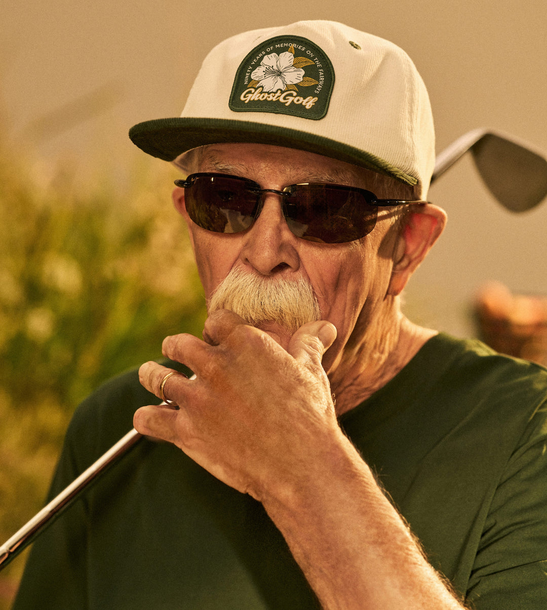 Senior Male model with his hadn on his mouth in a swaggy demeanor wearing a green tee and white hat holding golf club on his shoulder 