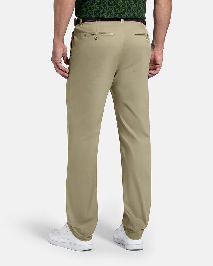 Back View of Sand Patrons Pleated Pant on white male model with green polo tucked in wearing brown belt and shoes #color_sand