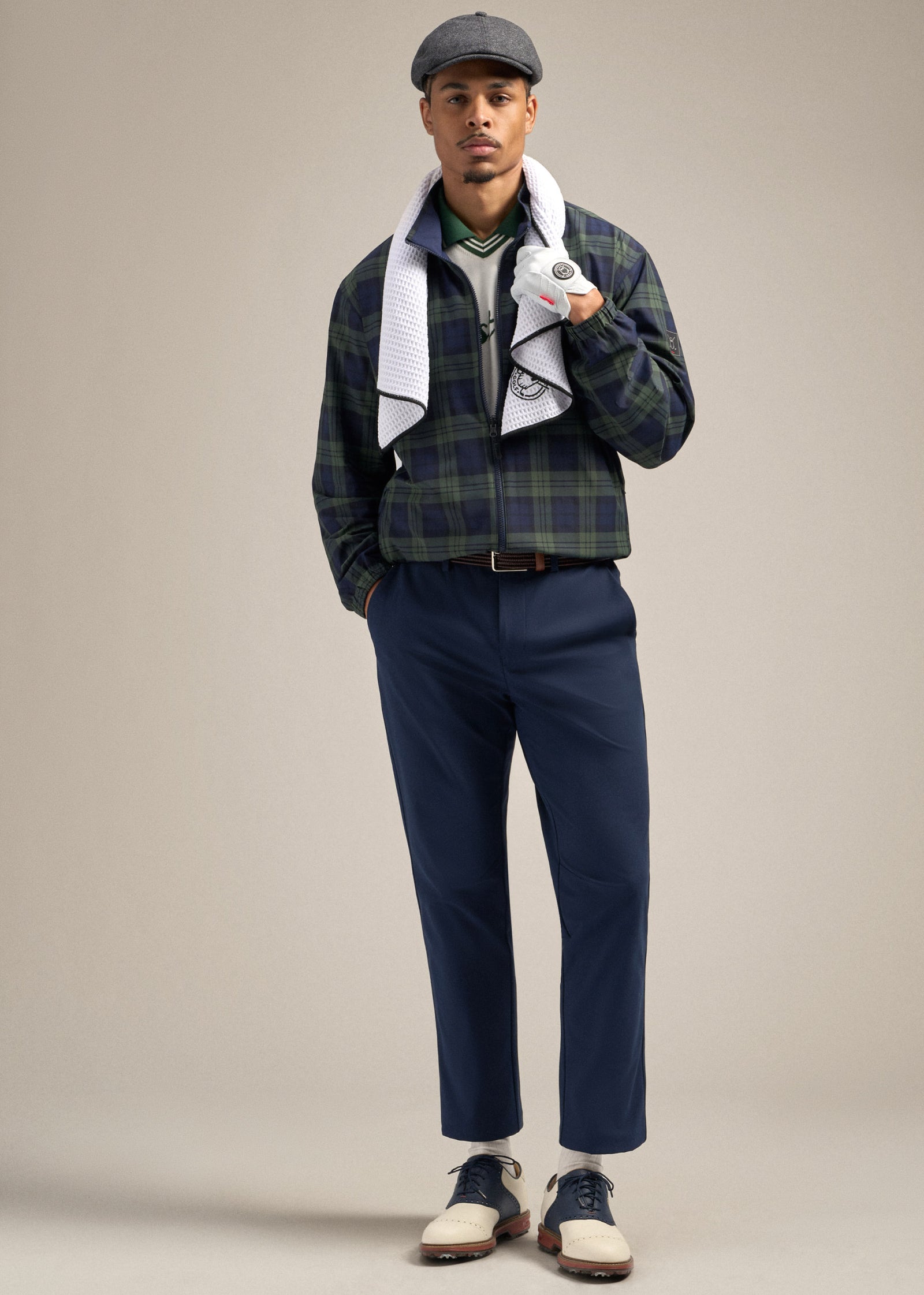 Full body shot of a Black male model wearing a flat cap and a reversible plaid spectator jacket. One hand is in his pocket while the other holds a golf towel draped around his neck. He is wearing navy pants paired with golf shoes, posed in a relaxed yet confident stance.