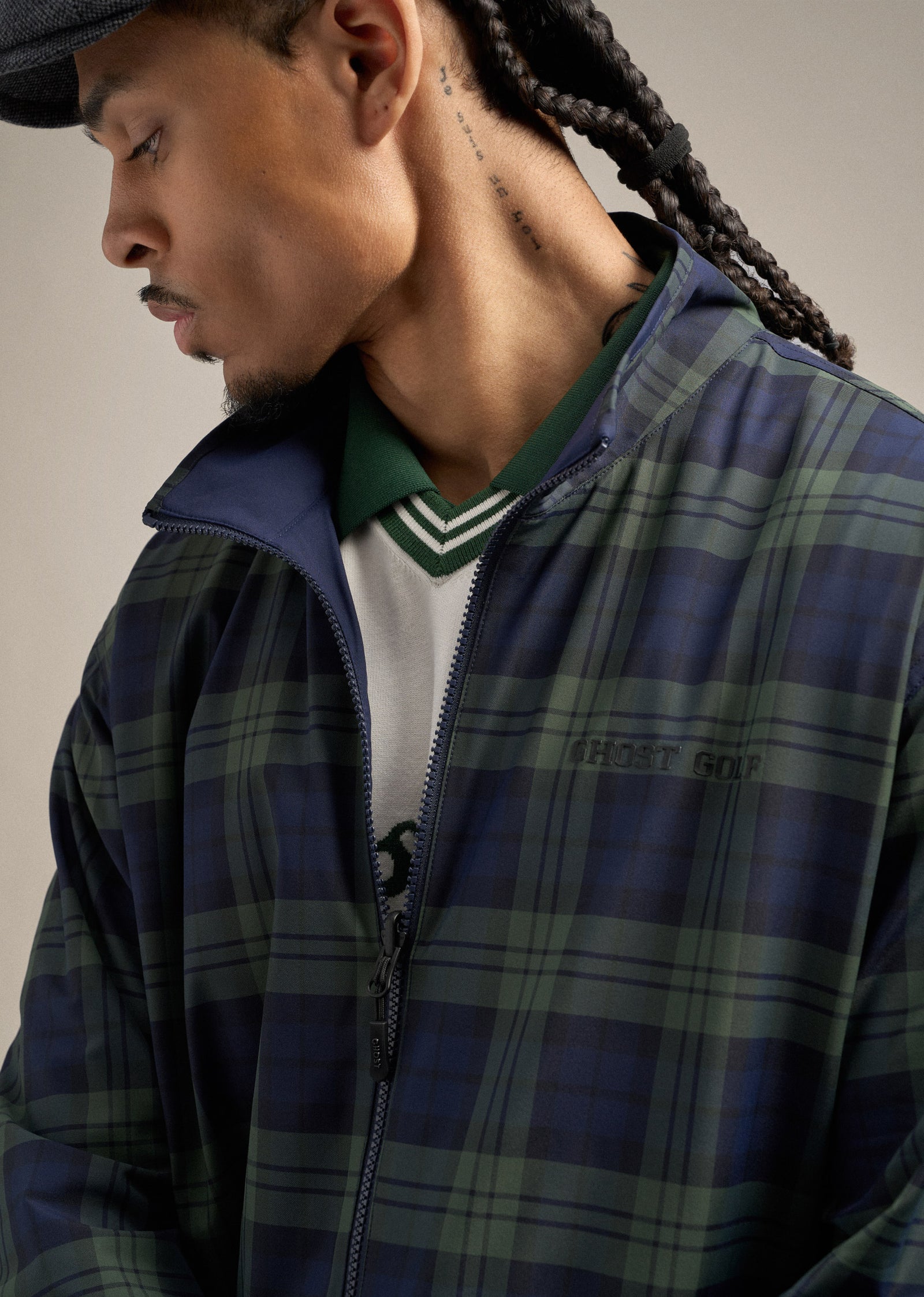 Close-up of a Black male model wearing a reversible plaid spectator jacket layered over a white sweater vest and a green polo. He is looking down toward his left, creating a refined and thoughtful pose.