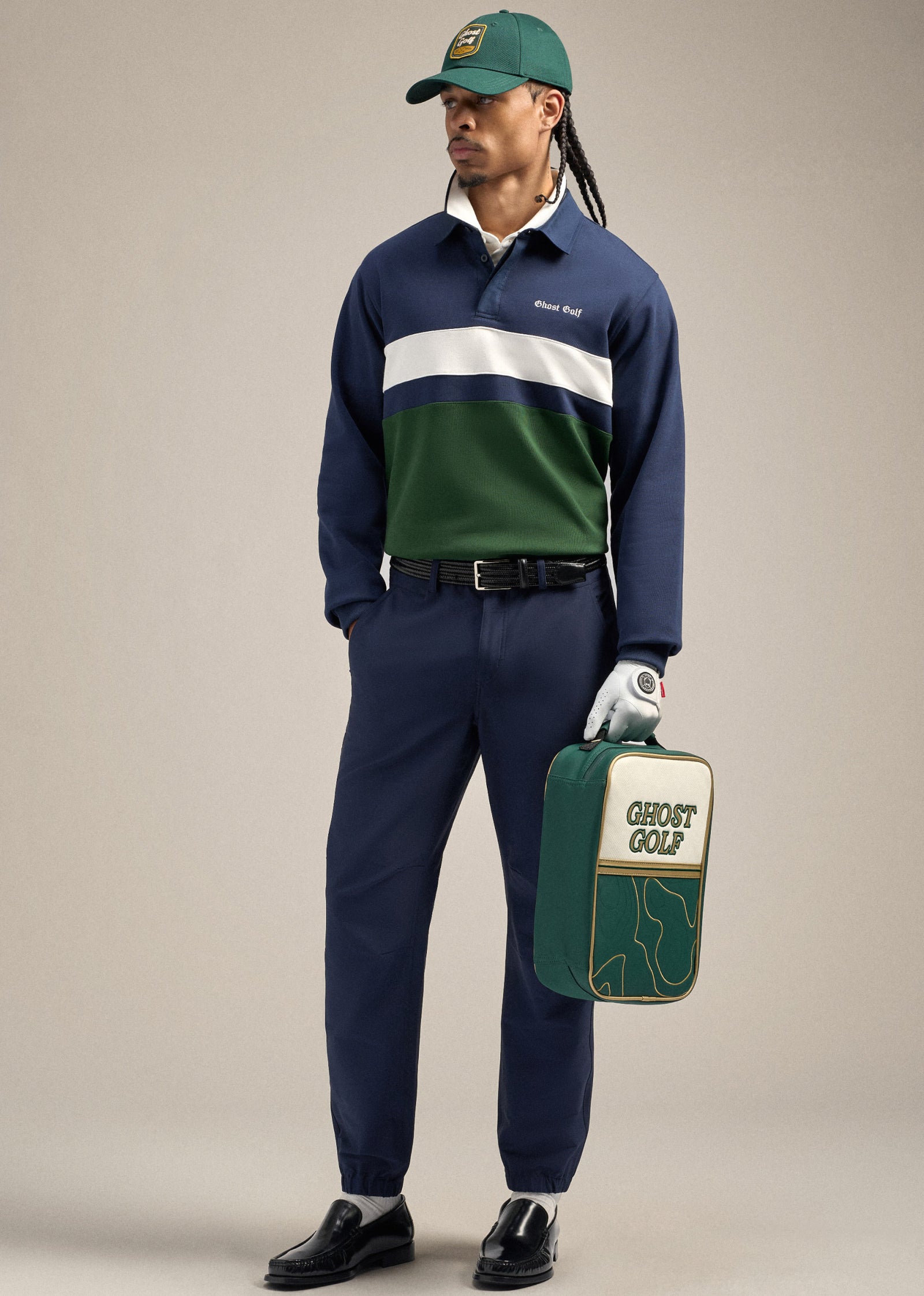 Full body shot of a Black male model wearing a navy polo sweatshirt tucked into navy pants with a brown belt. He is wearing dress shoes and a green golf hat. The model is posing with one hand in his pocket while holding a green shoe bag and a wearing a white golf glove in the other hand, looking toward his right.