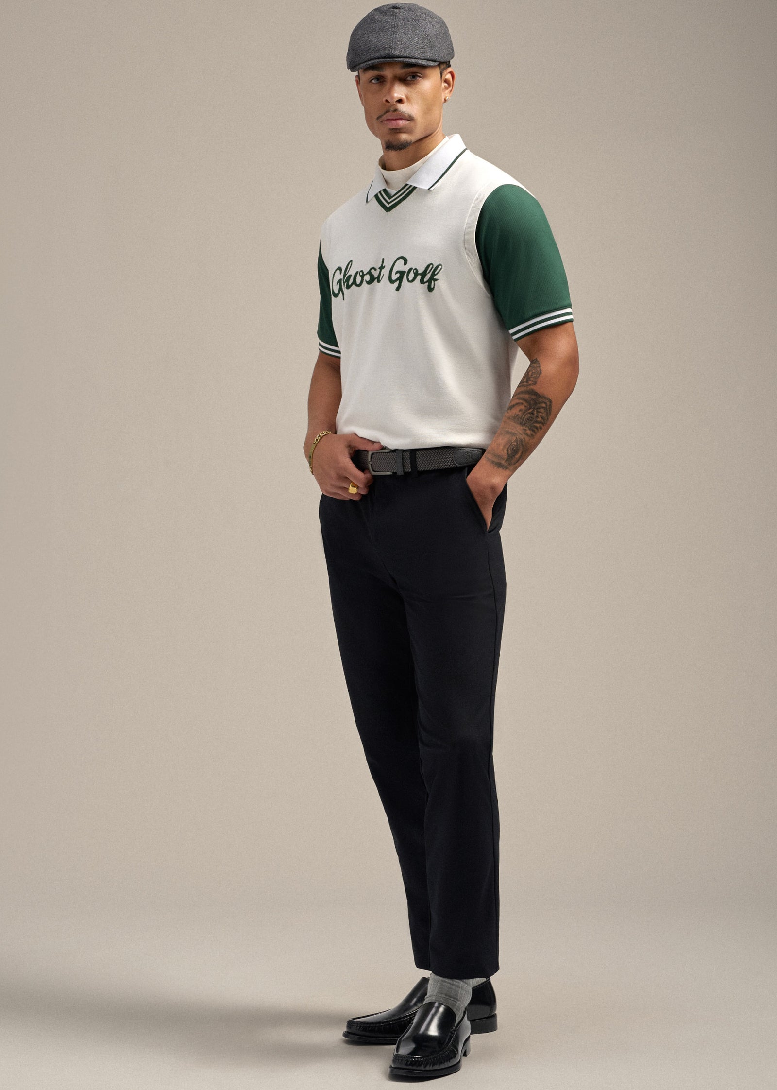 Full body shot of a Black male model posing elegantly. He is wearing a white sweater vest layered over a green polo shirt, paired with black pants. One hand is holding his golf belt while the other rests in his pocket. He is styled with a flat cap and simple, clean black dress shoes, creating a polished and refined look.