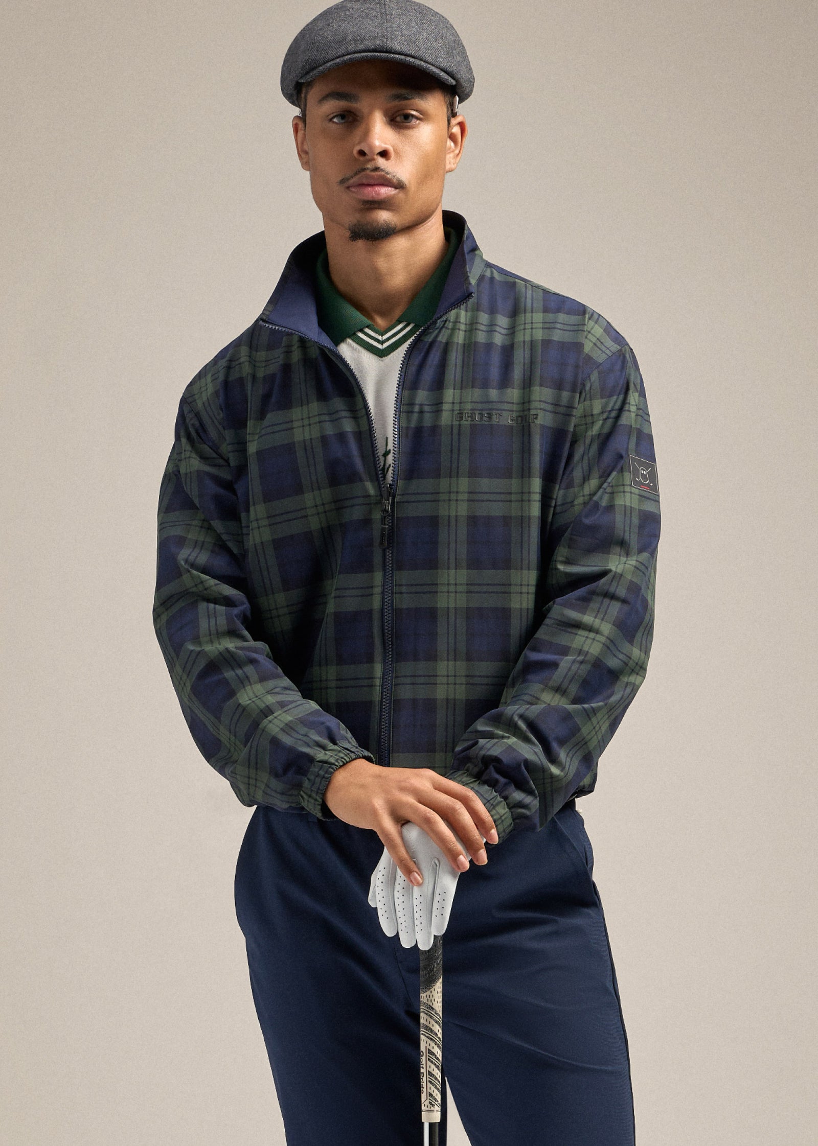 Front upper-body shot of a Black male model wearing a reversible plaid spectator jacket layered over a white sweater vest and a green polo, paired with navy pants. His hands are placed elegantly on top of a golf club—his gloved hand holding the club with the other hand resting on top. He is looking straight ahead, creating a refined and composed pose.