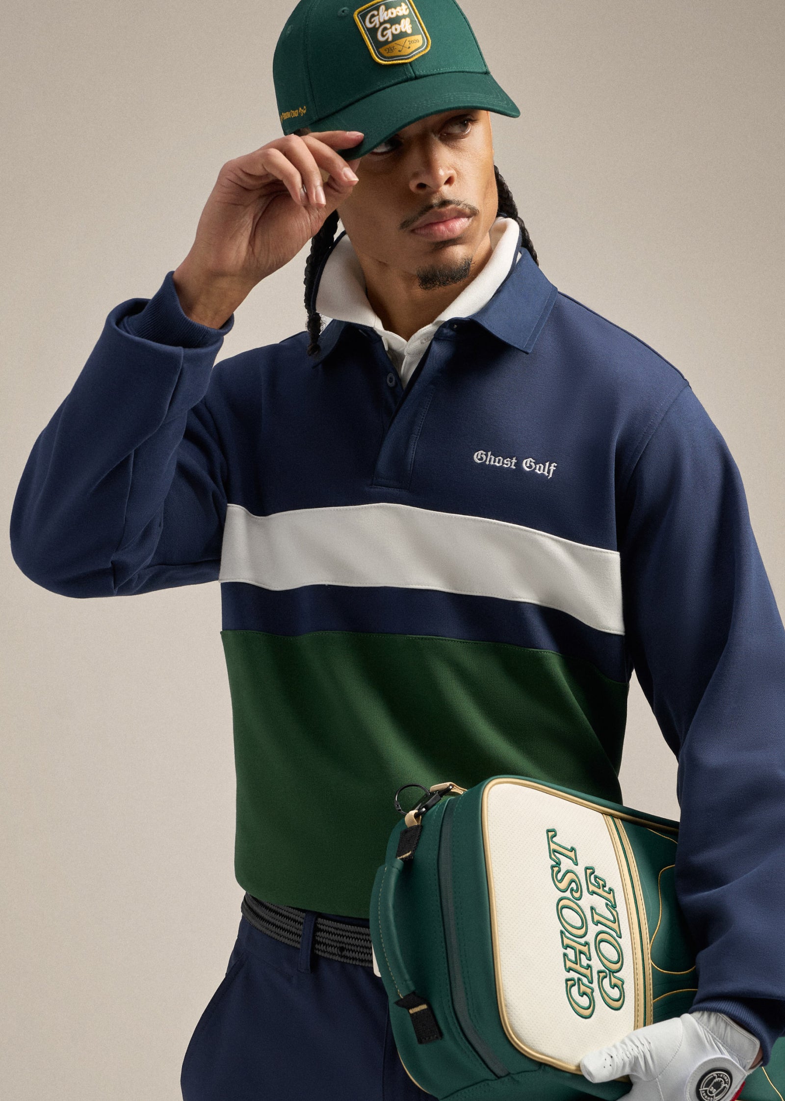 Close-up shot of a Black male model tilting his green golf hat with one hand while holding a green shoe bag with the other. He is wearing a navy polo sweatshirt and looking toward his left, creating a relaxed and confident pose.
