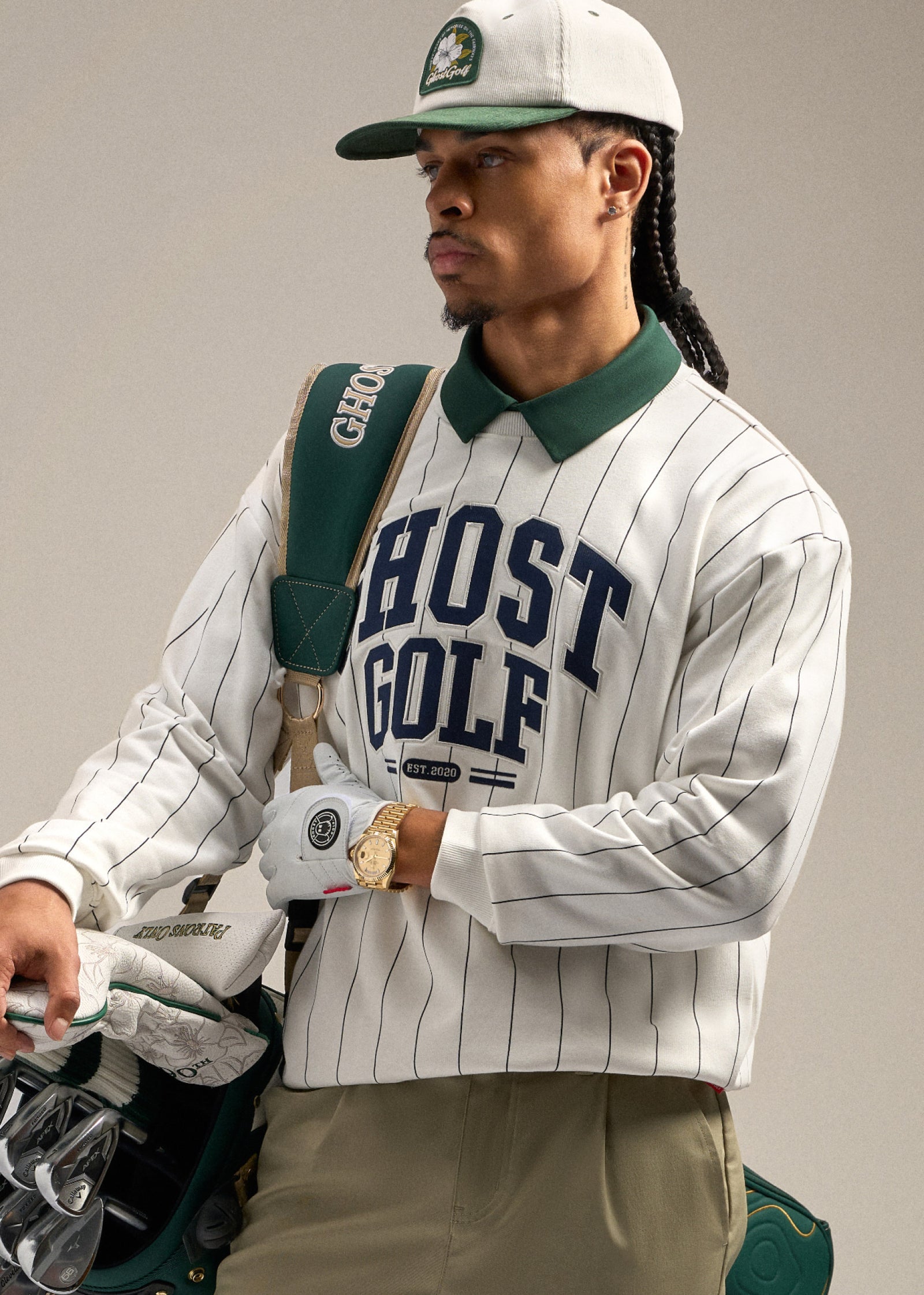 Black male model holding a golf bag over his shoulder, with his gloved hand securing it in place. He is wearing a white striped sweatshirt with Ghost Golf branding and a green polo underneath, paired with a white hat. He is looking toward his left, creating a confident golf-inspired pose.