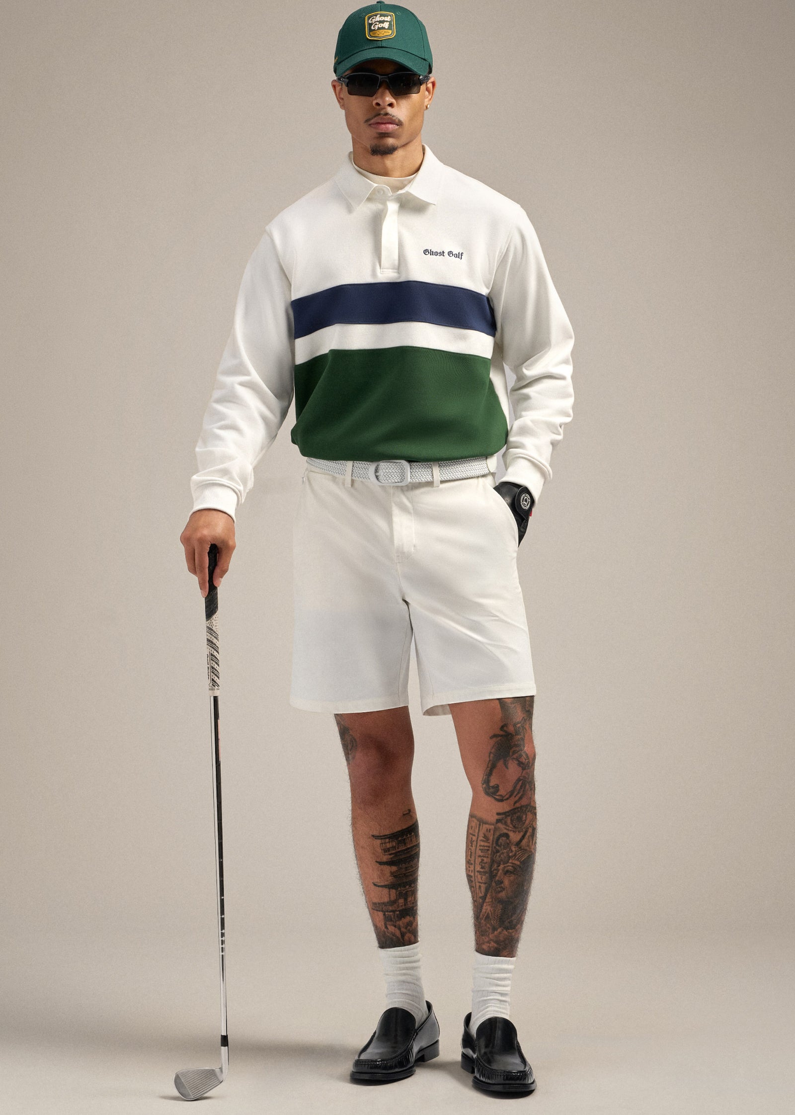 Full body shot of a Black male model wearing a white spectator polo sweatshirt paired with white shorts and a white belt. He is wearing a black glove, with his right hand in his pocket while the other hand holds a golf club resting on the floor. He is styled with a green cap, black dress shoes, and sunglasses, creating a confident and polished golf-inspired pose.