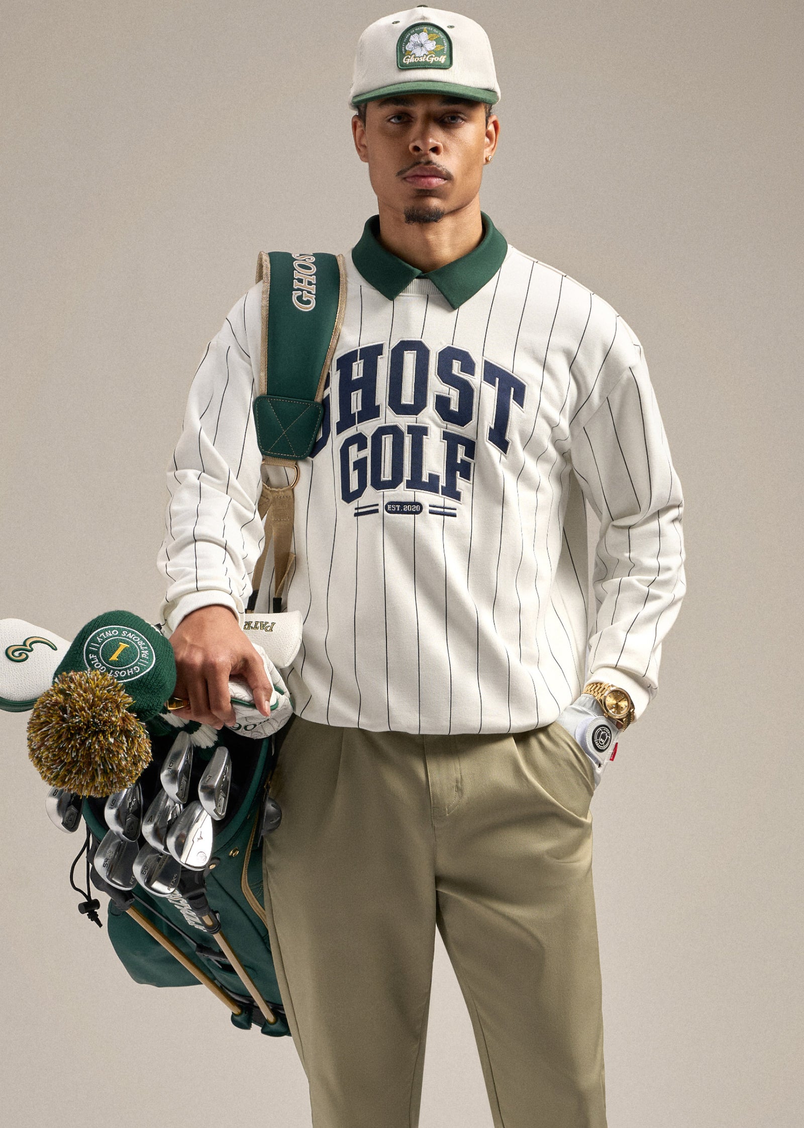 Black male model holding a golf bag over his shoulder, with his gloved hand in his pocket. He is wearing a white striped sweatshirt with Ghost Golf branding over a green polo, paired with sand wedge–colored pants and a white hat. He is looking straight ahead, creating a clean and confident golf-inspired pose.