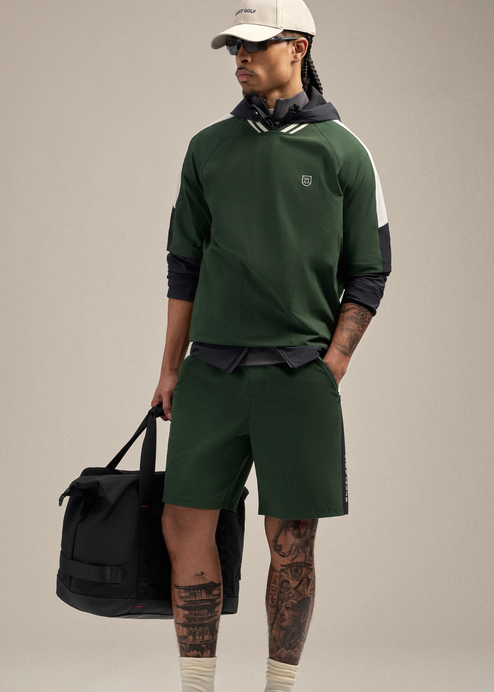 Front shot of a Black male model looking toward his left, wearing a beige hat and sunglasses. He is dressed in a green fairway windshirt and matching windshorts, with one hand in his pocket and the other holding a black duffle bag, creating a clean and sporty golf-inspired look.