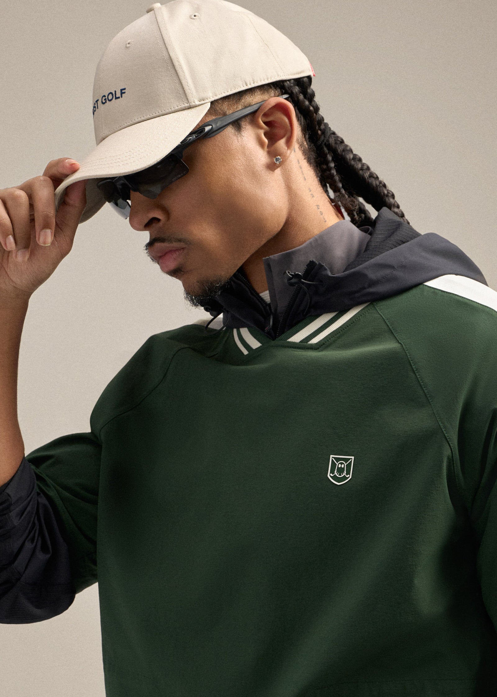 Close-up shot of a Black male model looking toward his left while tilting his hat with one hand. He is wearing sunglasses and a green fairway windshirt featuring a Ghost icon badge on the garment, creating a clean and sporty golf-inspired look.