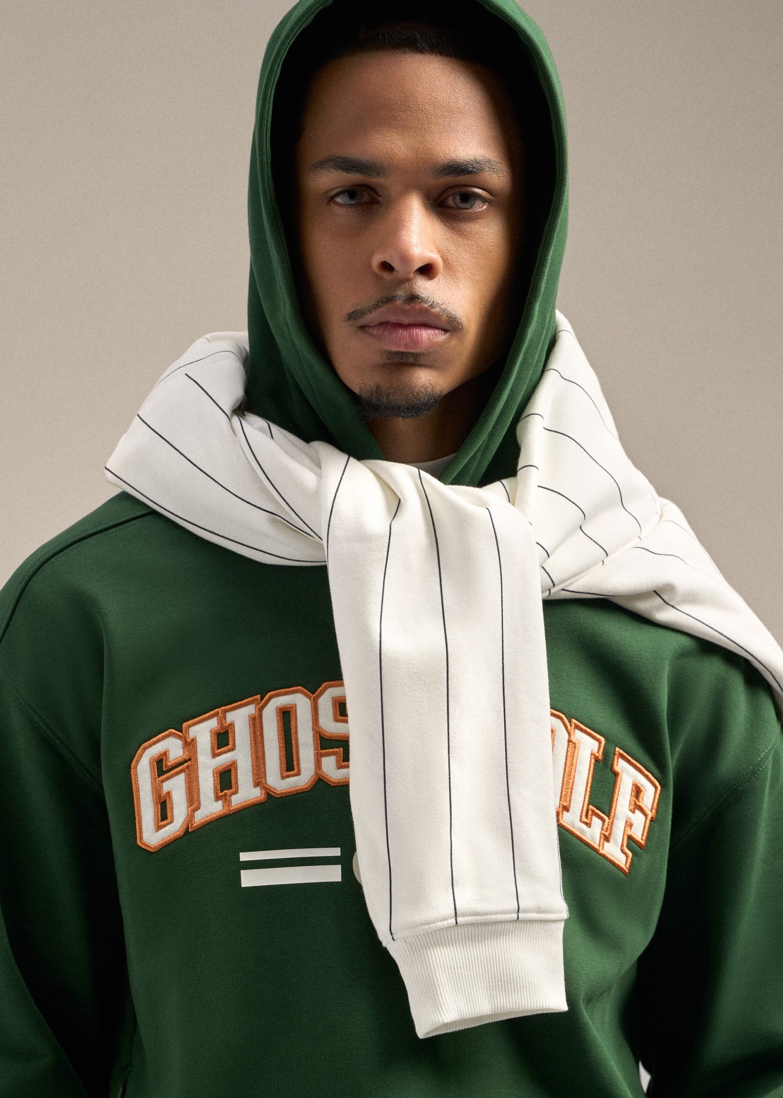 Black male model wearing a green hoodie with a white striped sweatshirt wrapped around the neck and shoulder area, creating a relaxed and layered look.