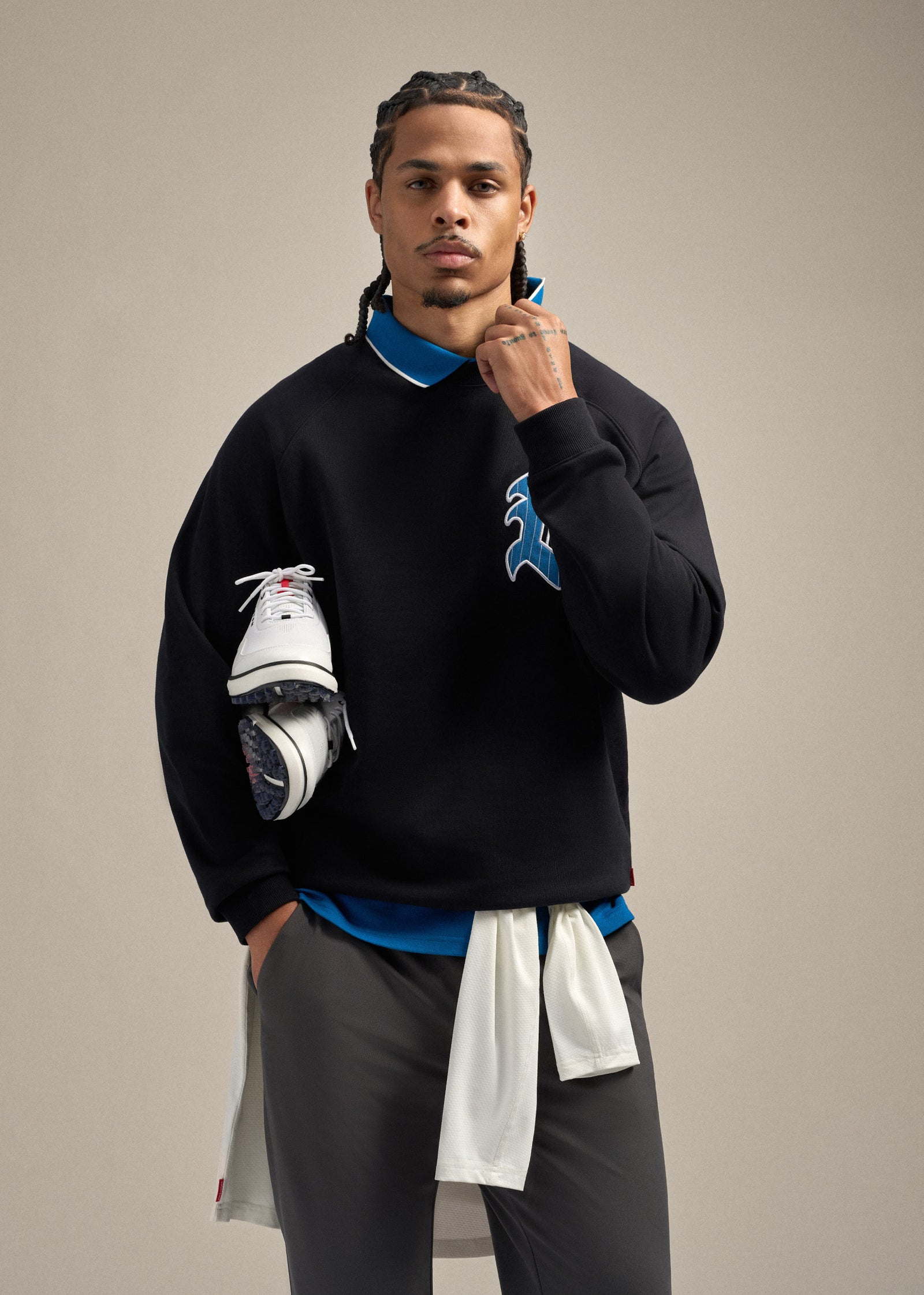 Black male model wearing a black sweatshirt with a large G hit on the left chest, layered over a royal blue polo. He is wearing grey pants and looking straight ahead. One hand is holding the collar while the other arm carries golf shoes, with that hand resting in his pocket, creating a confident and styled golf-inspired pose.