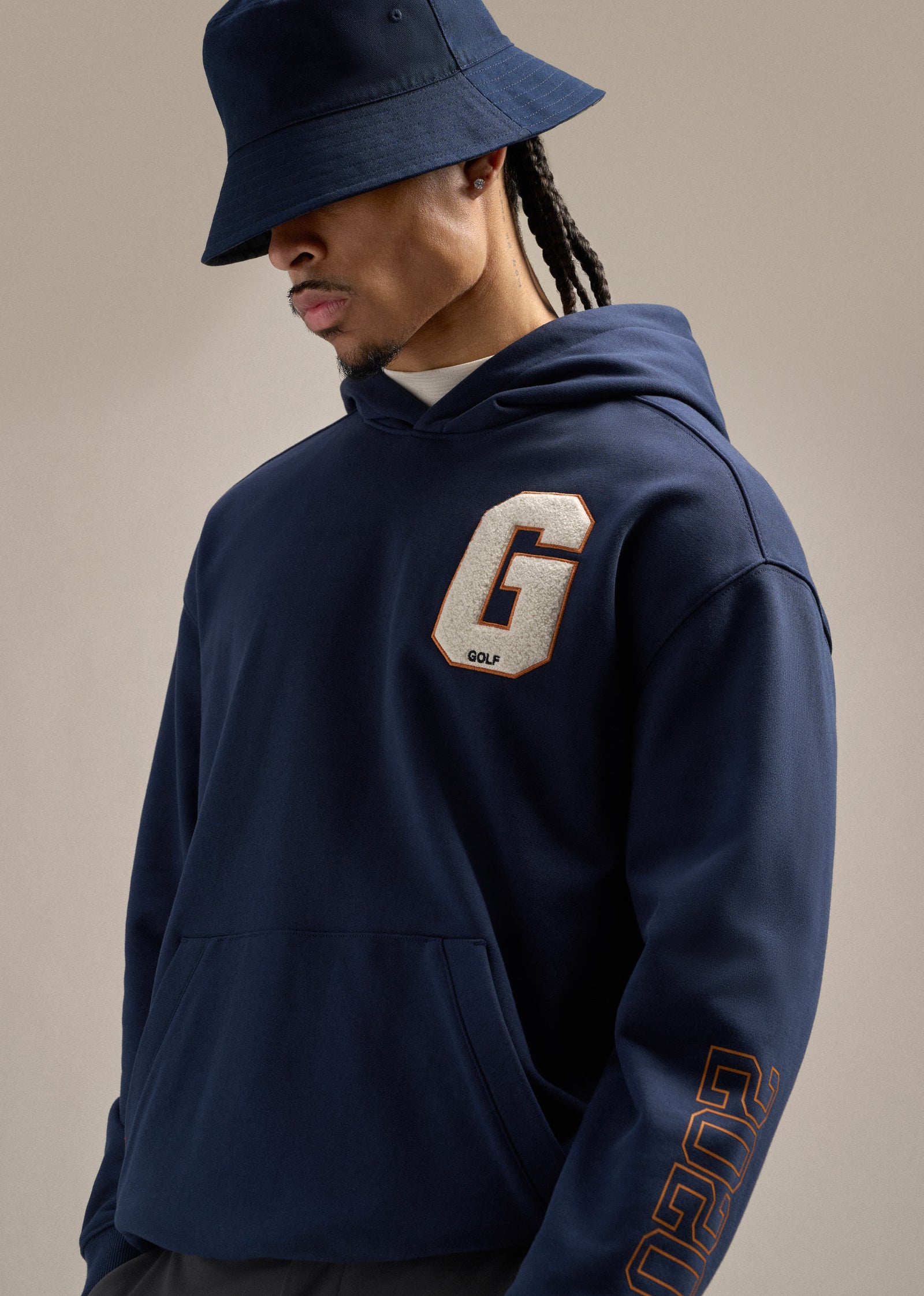 Black male model wearing a navy hoodie with a large G hit on the left chest, paired with a navy reversible bucket hat. He is looking toward his left, creating a relaxed and modern golf-inspired look.