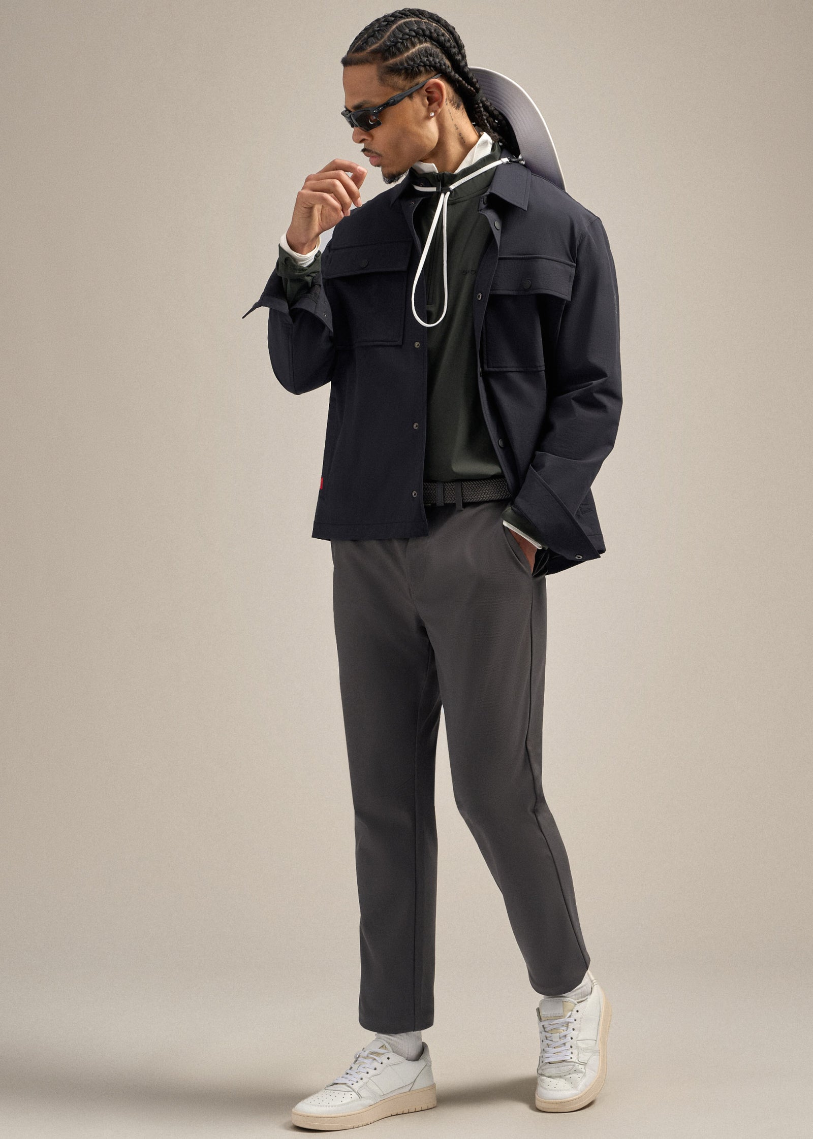 Black male model wearing a black jacket, grey pants, green mock jacket and a polo. One hand is in his pocket while he faces toward his left. His other hand is near his face, with a boonie hat draped on his back, creating a relaxed and stylish pose.