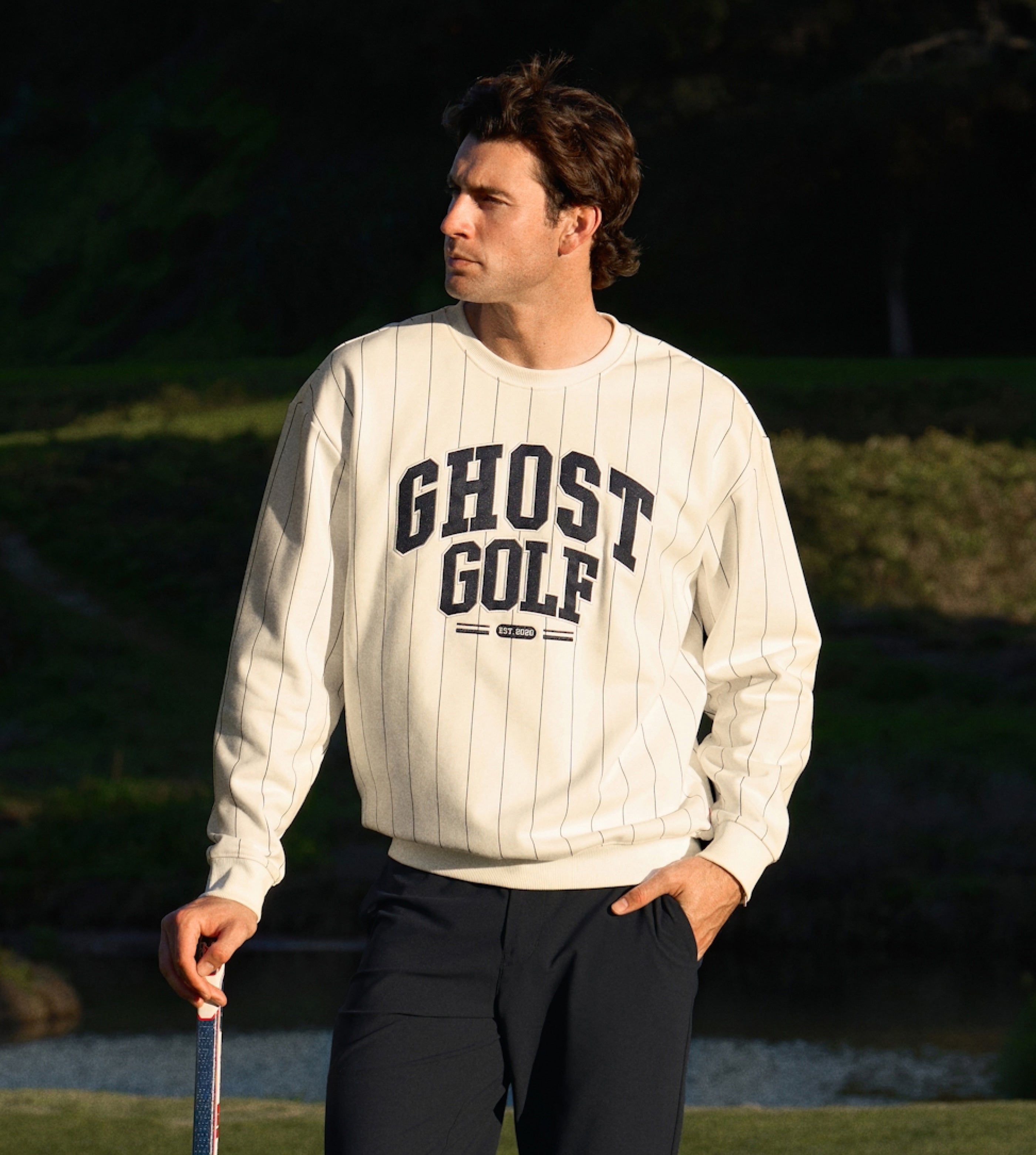 White male golfer on course in a white Ghost Golf sweatshirt and black pants, hand in pocket.