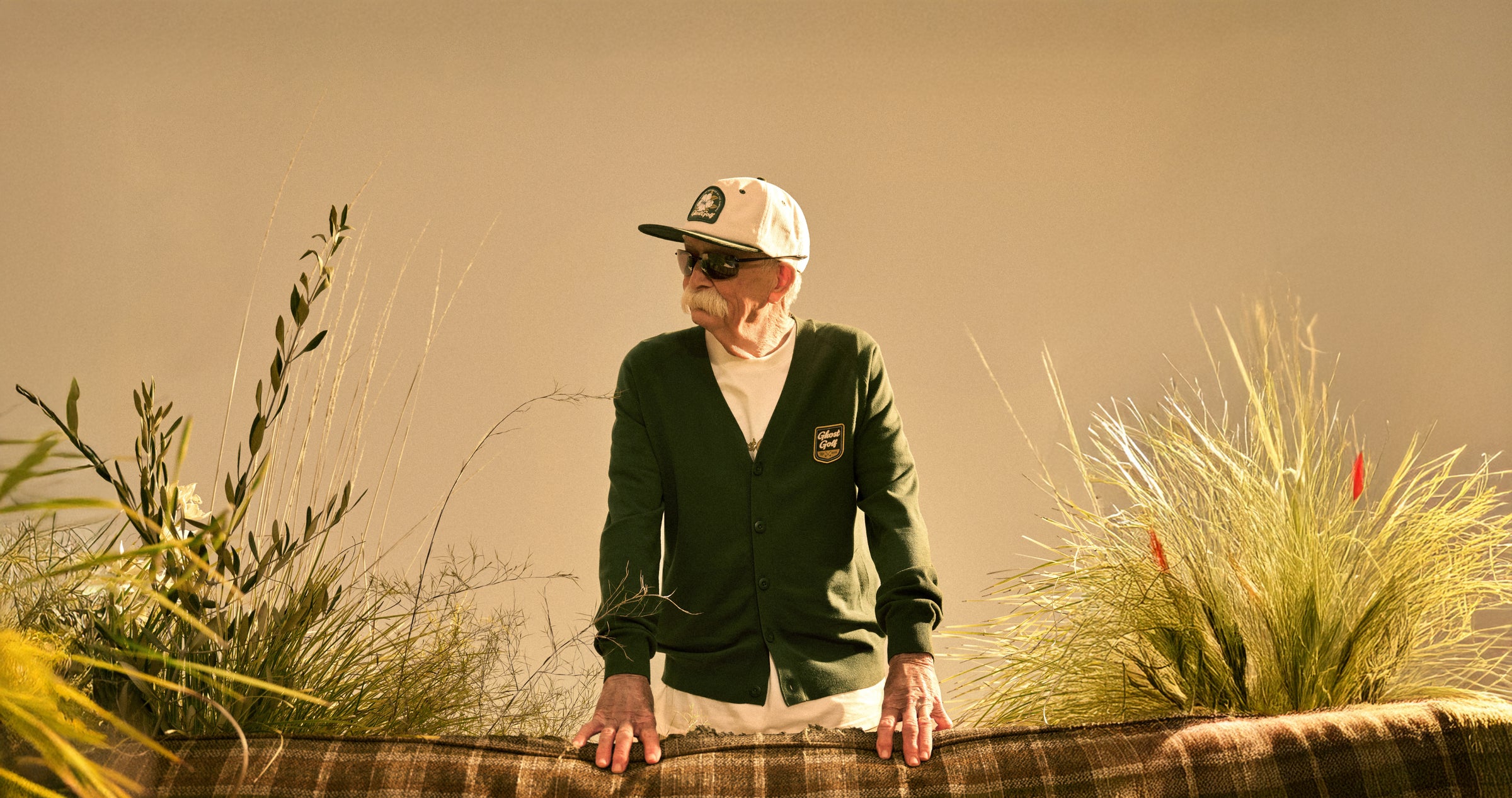 Senior male model standing behind a couch in a beige studio setting, hands resting on the back of the couch. He is looking to his right and wearing a white hat, sunglasses, and a green cardigan.