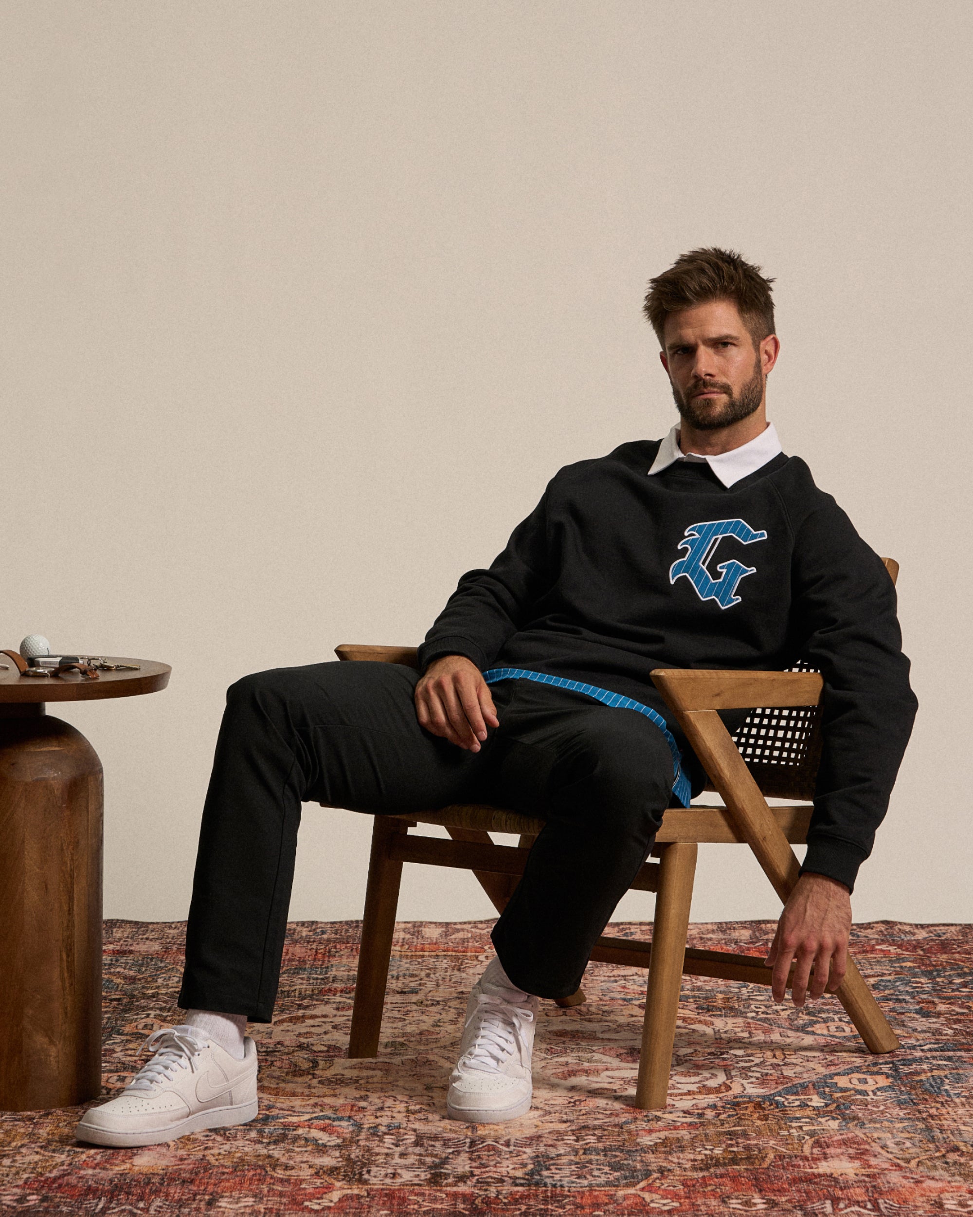 Male model seated on a chair in a studio setting, leaning forward in a relaxed, confident pose. He is wearing a black sweatshirt with a ‘G’ logo on the chest and black pants.
