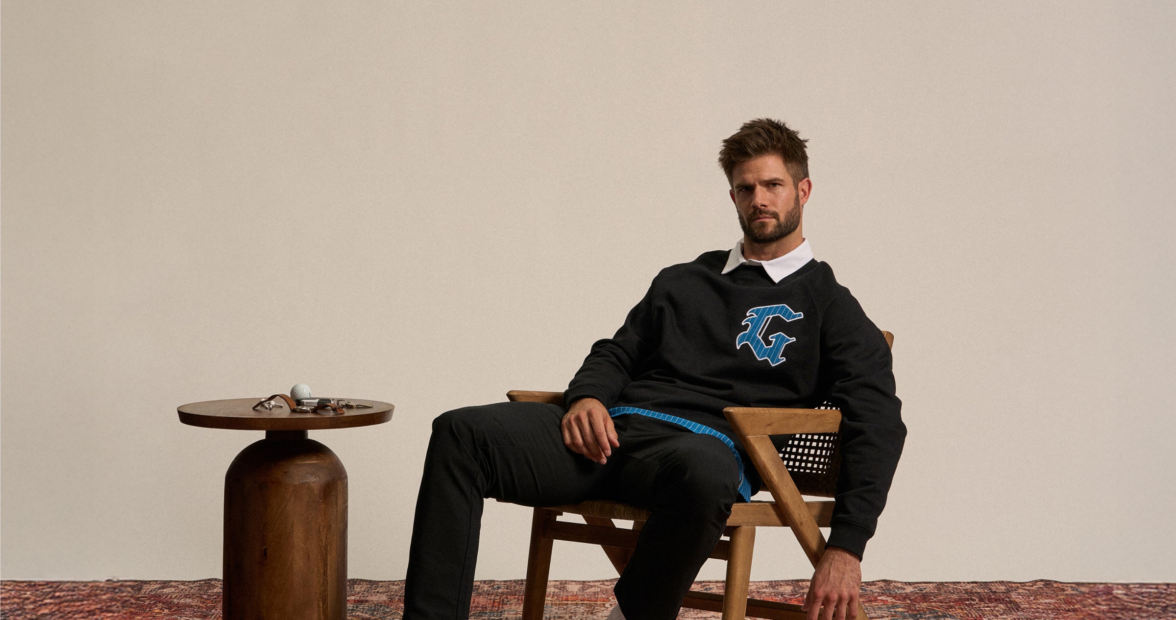 Male model seated on a chair in a studio setting, leaning forward in a relaxed, confident pose. He is wearing a black sweatshirt with a ‘G’ logo on the chest and black pants.