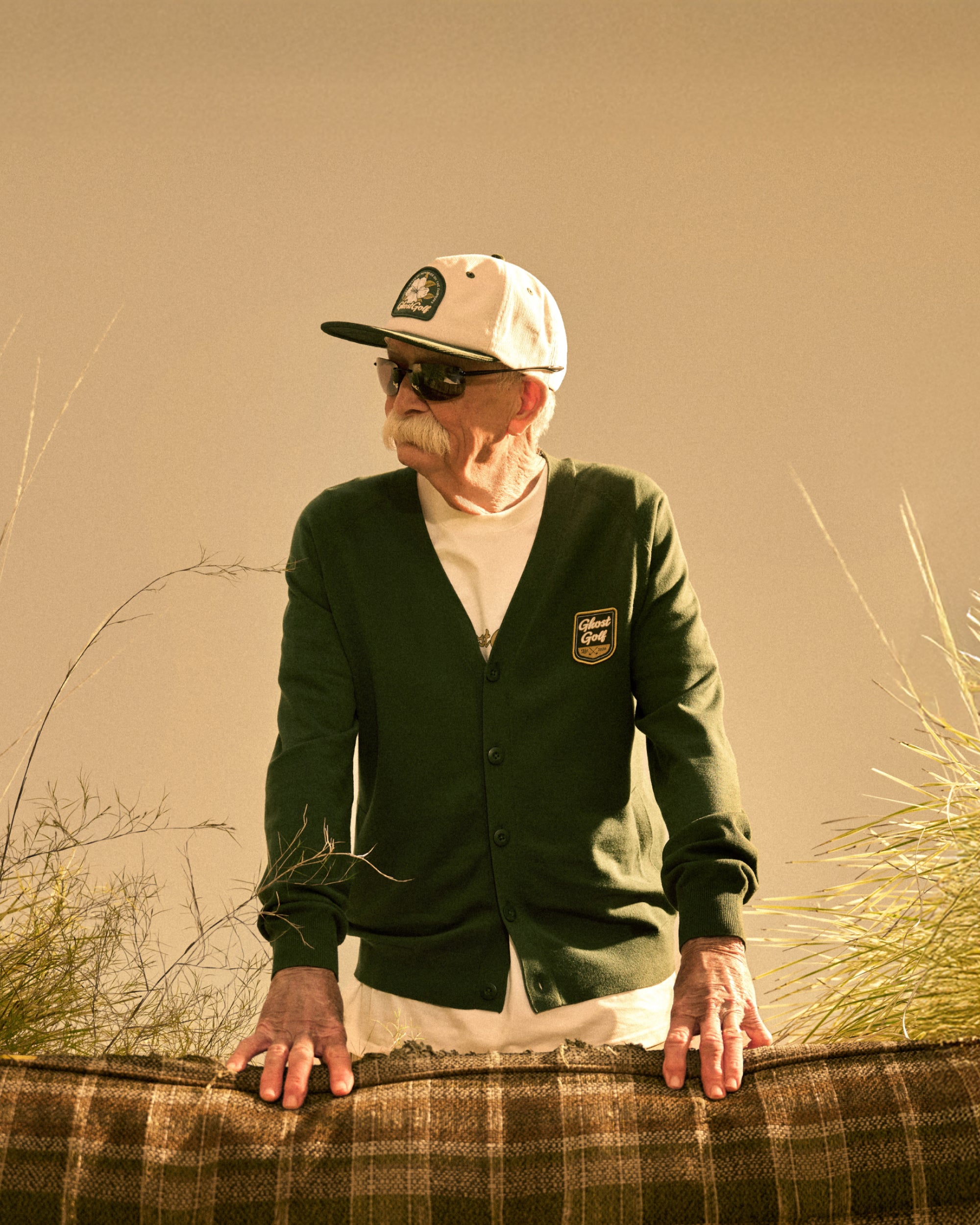 Senior male model standing behind a couch in a beige studio setting, hands resting on the back of the couch. He is looking to his right and wearing a white hat, sunglasses, and a green cardigan.