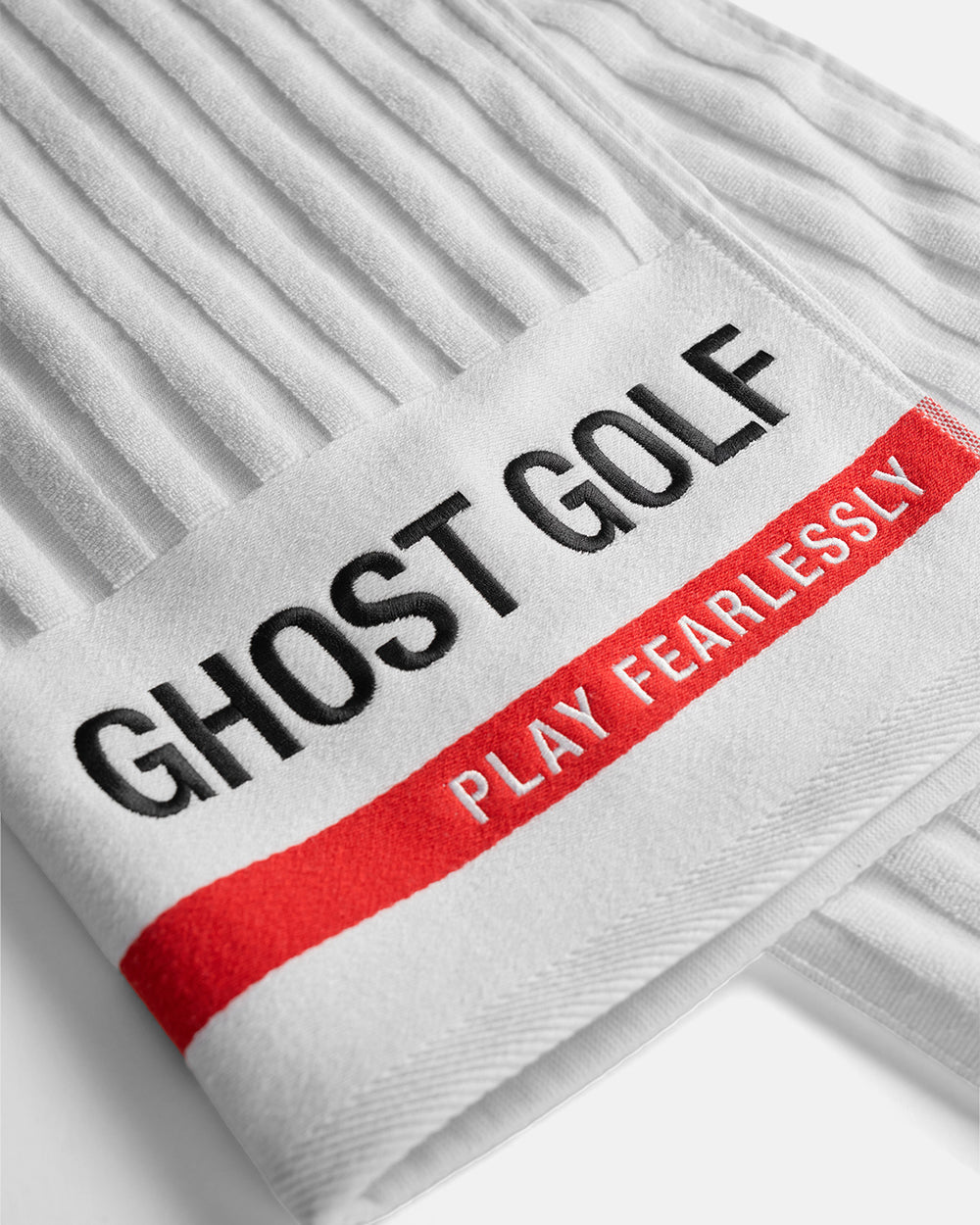 Closeup Product shot of White Tour Pro Caddie Towel folded flat, featuring the Ghost Golf logo on the bottom right, a red stripe across the bottom, and “Play Fearlessly” text #color_white