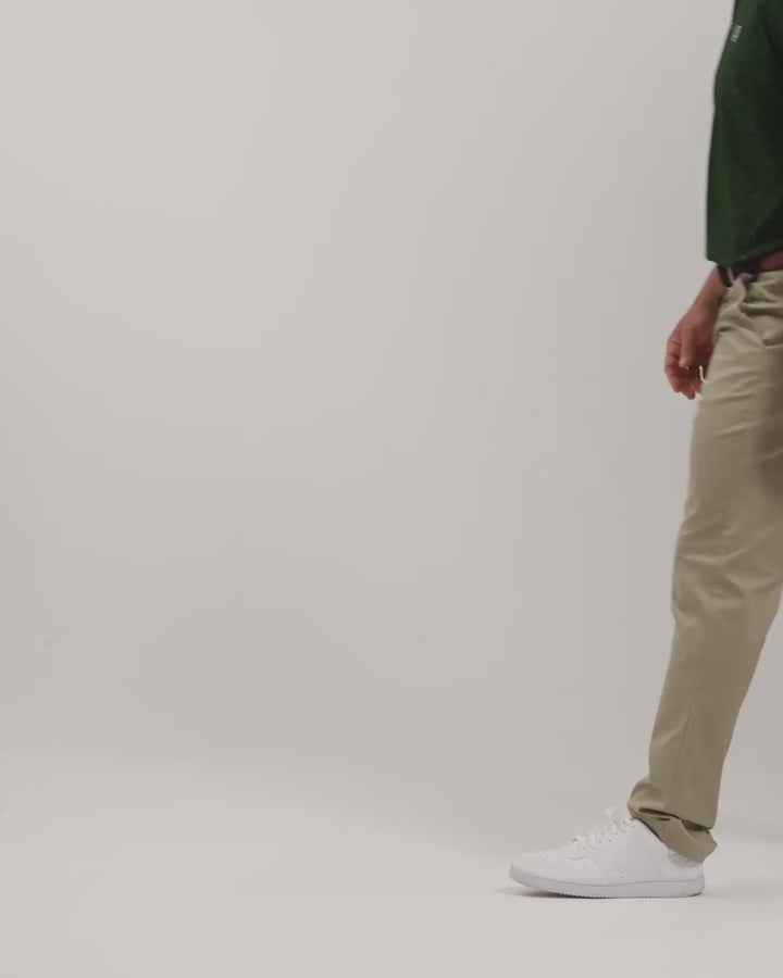 Male model walking naturally showcasing Sand Patrons Pleated Pants from all angles wearing green polo and white shoes #color_sand-wedge