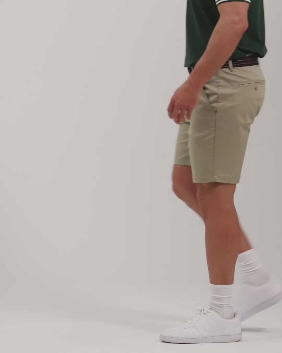 Male model walking naturally showcasing Sand Patrons Pleated Shorts from all angles wearing green polo and white shoes #color_sand-wedge