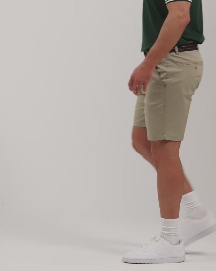 Male model walking naturally showcasing Sand Patrons Pleated Shorts from all angles wearing green polo and white shoes #color_sand-wedge
