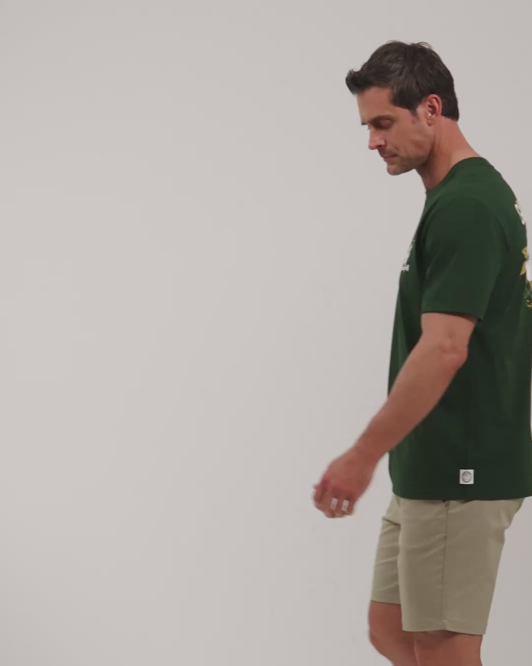 Male model walking naturally showcasing Bogey Green Patrons Azalea Tee from all angles wearing beige shorts and white shoes #color_bogey-green